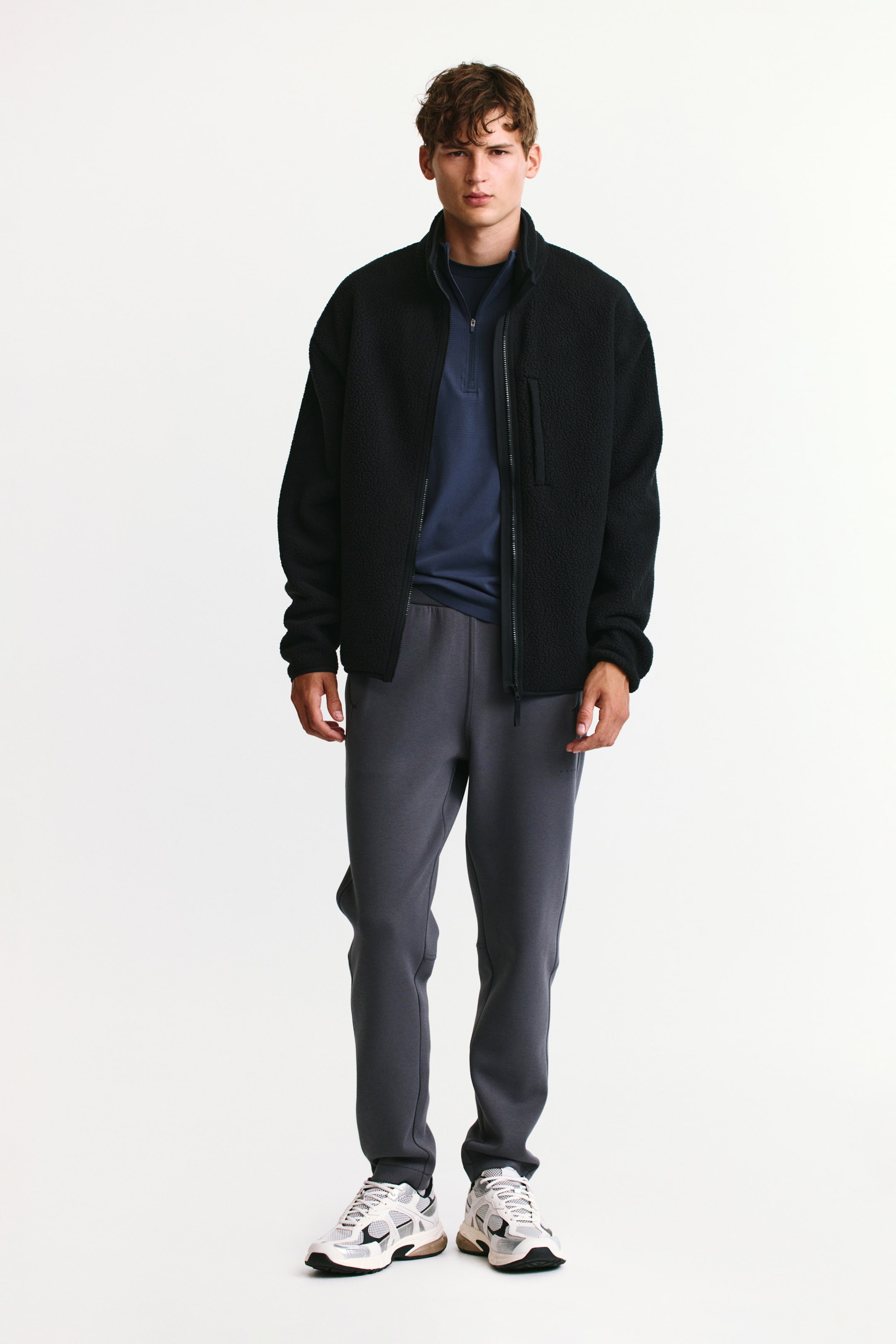 View larger image: A black fleece jacket with a high collar and a zippered chest pocket is being worn open by the model. The jacket is layered over a dark blue quarter-zip pullover and paired with grey sweatpants and light-coloured sneakers.