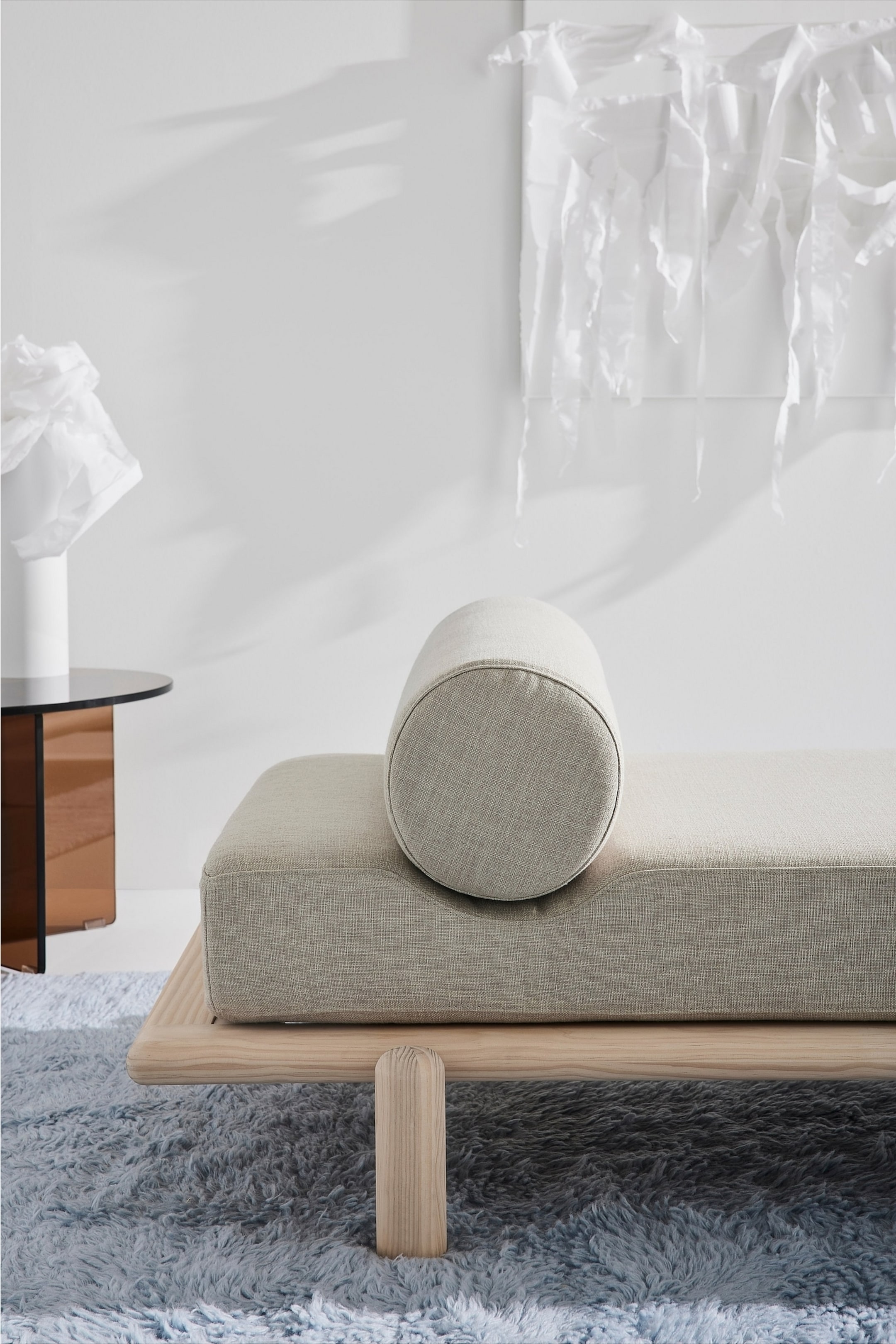 Visa större bild: En ljusbeige schäslong är placerad i en vinkel från framsidan, med en rektangulär kudde i texturerat tyg och en subtil fördjupning. En matchande cylindrisk valuavalkudde vilar på denna fördjupning. Schäslongen stöds av en ljus trästomme med en synlig, avrundad träben.