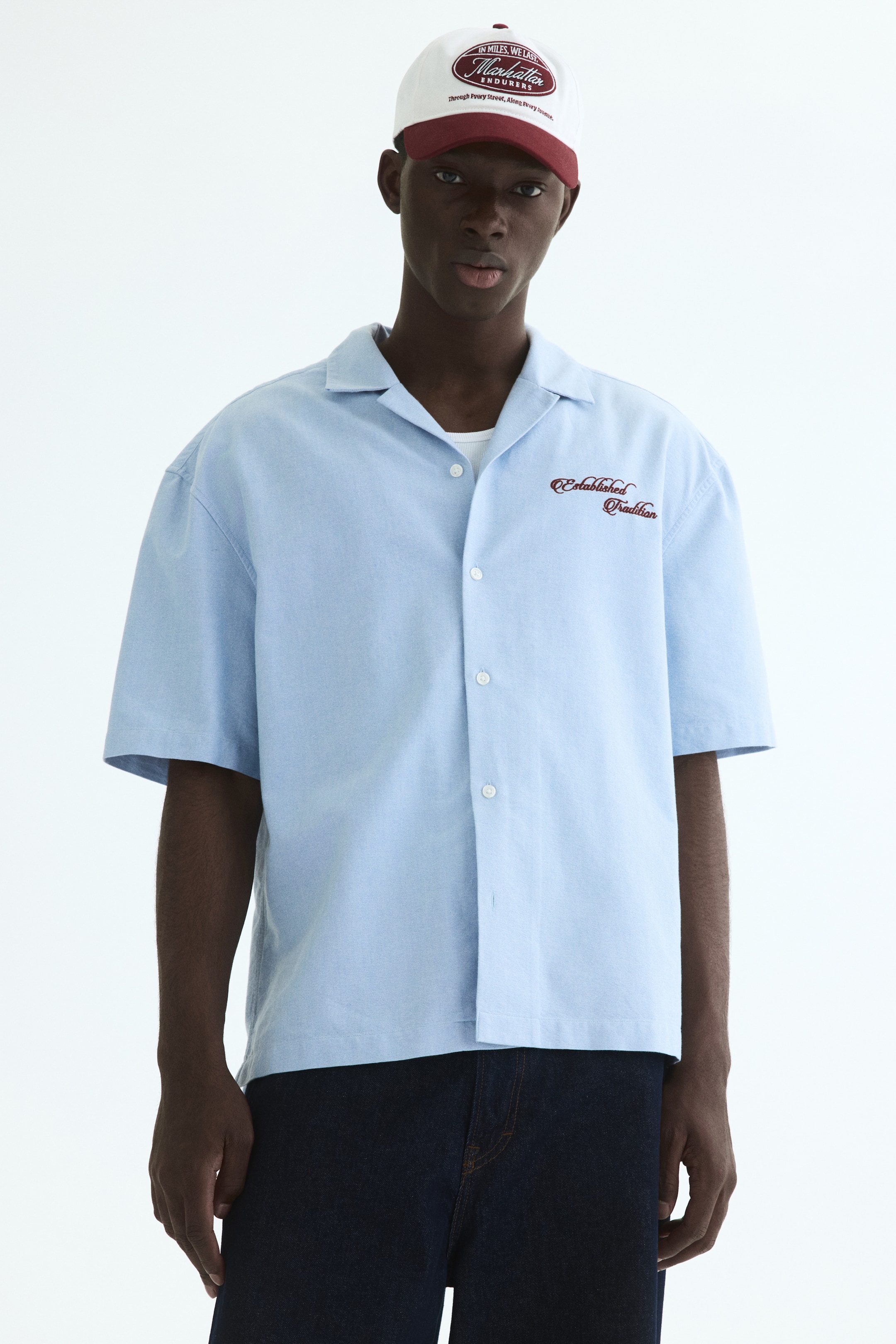 View larger image: The light blue short-sleeved shirt features a camp collar and a button-up front with white buttons. It displays "Established Tradition" embroidered in dark red on the left chest, and a white undershirt is visible beneath its open collar.