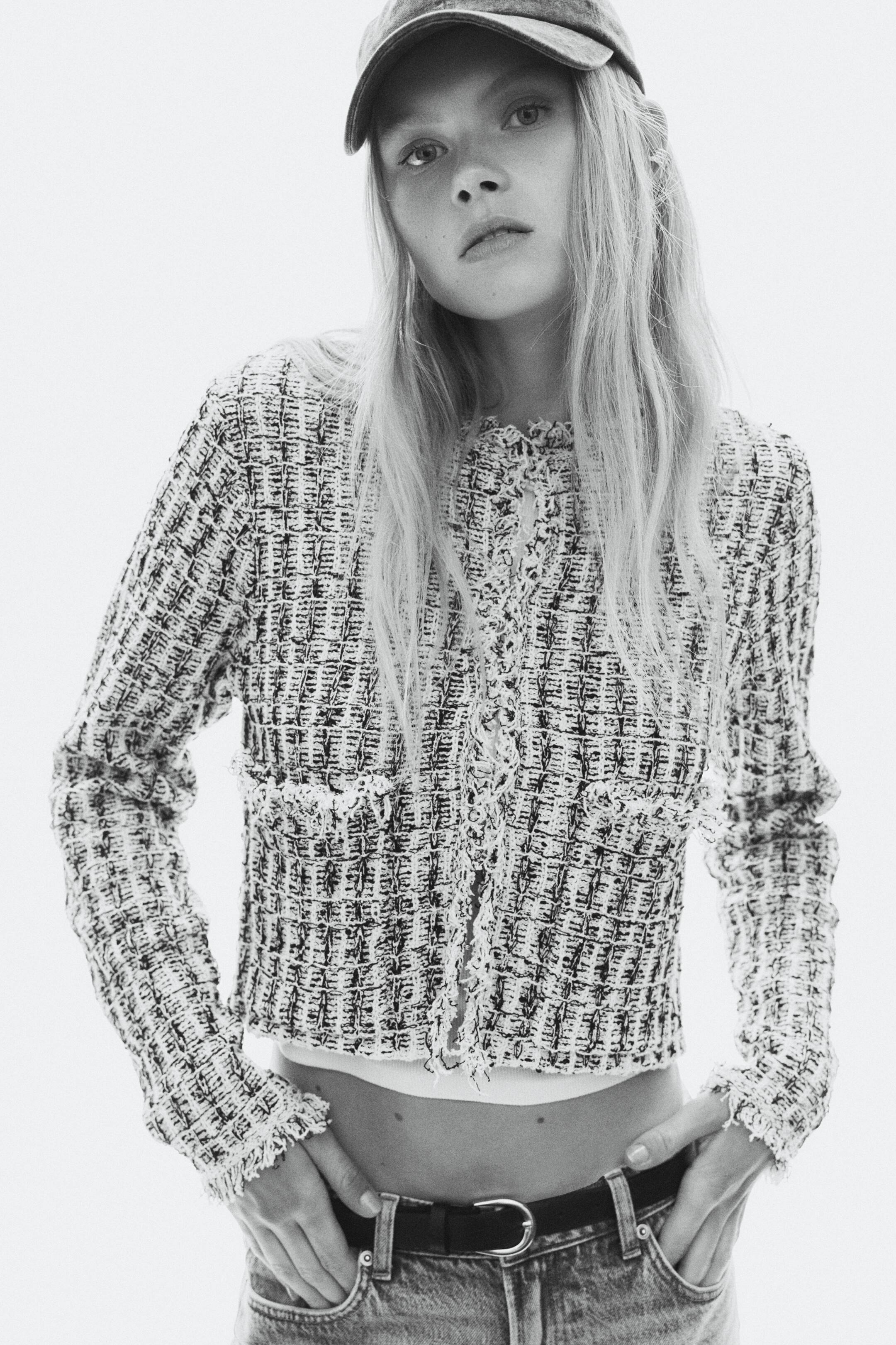 View larger image: The model is wearing a textured cropped tweed jacket with frayed trims along the collar, front opening, cuffs, and hem. The long-sleeved jacket is styled open over a white top and paired with low-rise jeans, a dark belt, and a baseball cap.