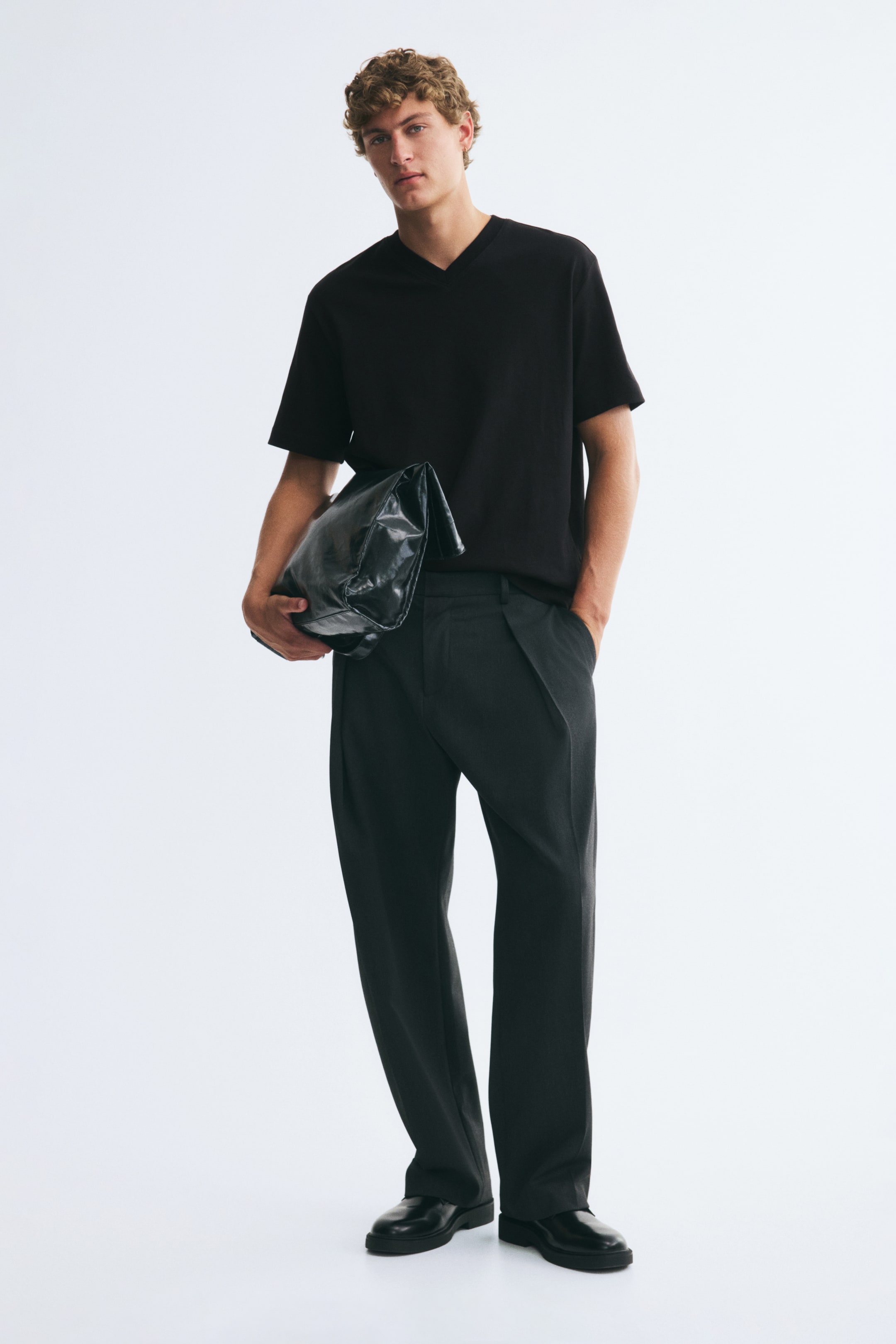 Xem ảnh lớn hơn: Áo thun jersey cotton interlock màu đen với cổ chữ V gân, tay ngắn và lai thẳng đang được người mẫu mặc. Áo được phối cùng quần tối màu dáng rộng và một chiếc túi đen được kẹp dưới một tay.