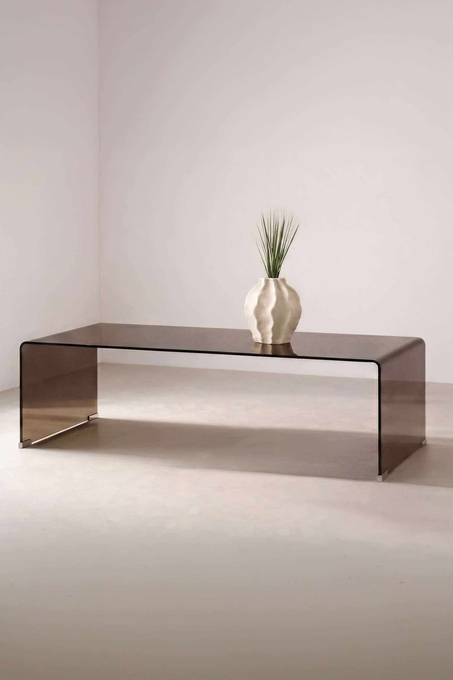 De Telemark Sofa Table toont een minimalistisch ontwerp uit Ã©Ã©n stuk van gebogen glas met een doorschijnende gerooktbruine afwerking. Het heeft een lang, plat tafelblad dat naadloos overloopt in watervalvormige zijdelen, die rusten op kleine metalen beschermende glijders.