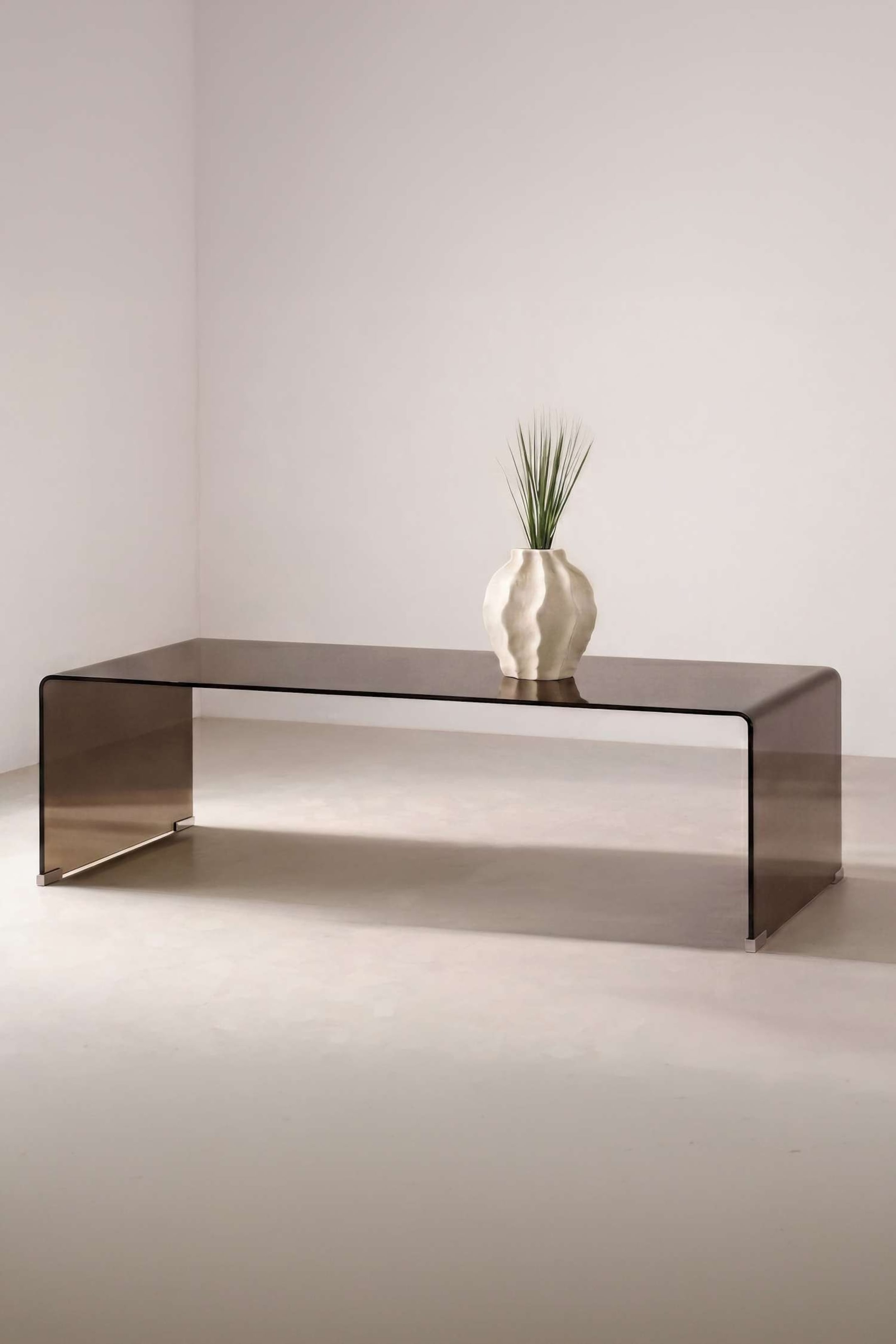 Ver imagen más grande: La mesa auxiliar Telemark exhibe un diseño minimalista en cascada, elaborado a partir de una sola pieza de vidrio ahumado translúcido, que se curva sin interrupción en cada extremo para formar las patas. En un ángulo que destaca su estructura larga y rectangular, la mesa sostiene sobre su superficie reflectante un jarrón de cerámica blanca texturizada con follaje verde.