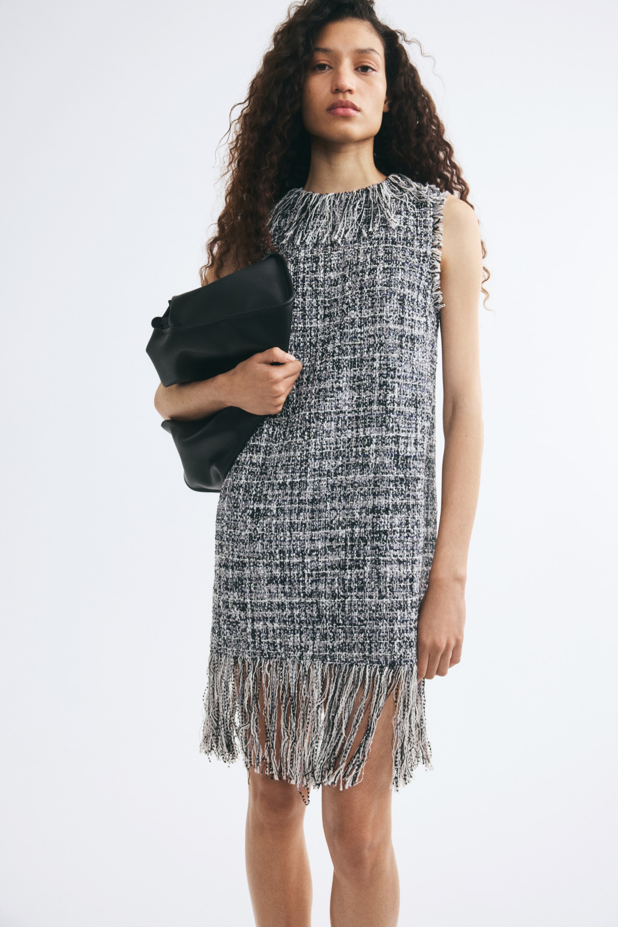 Voir une image plus grande: La mannequin porte une robe sans manches en tweed noir et blanc, avec un col montant à franges et un ourlet assorti à franges. Une grande pochette en cuir noir est tenue sous le bras de la mannequin.