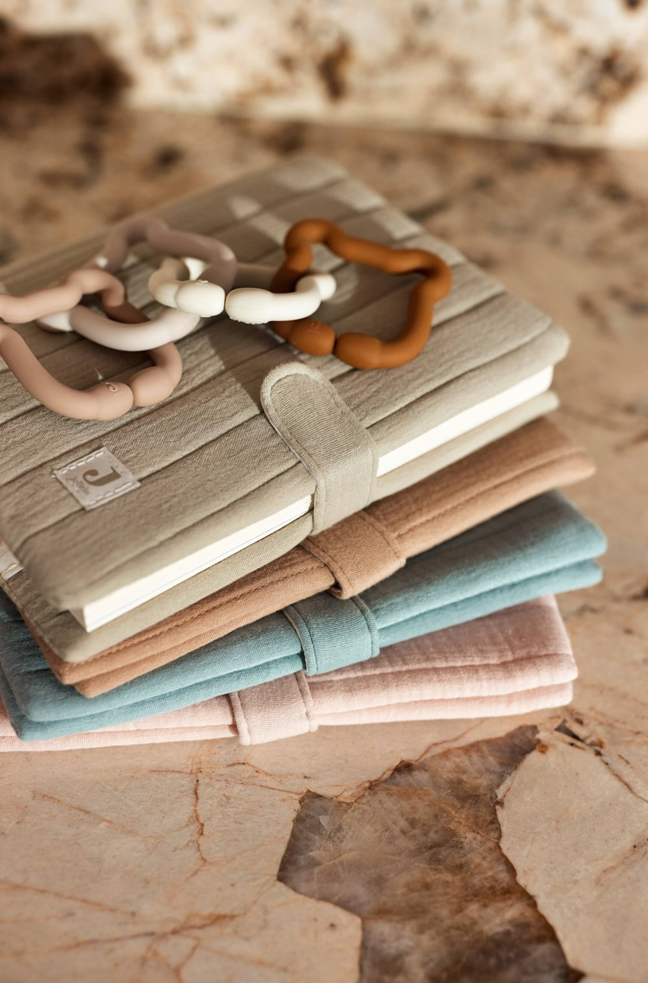 Agrandir l'image: Une pile de quatre sacs de rangement en tissu matelassé, dans des tons vert olive, beige, bleu poudré et rose pâle, est présentée en vue en plongée à trois quarts, chaque sac étant doté d’un matelassage vertical et d’une fermeture à sangle en tissu assortie. Des anneaux de jouets en silicone entrelacés, dans des tons terre atténués, reposent sur le sac vert, mettant en valeur sa structure douce et rembourrée.