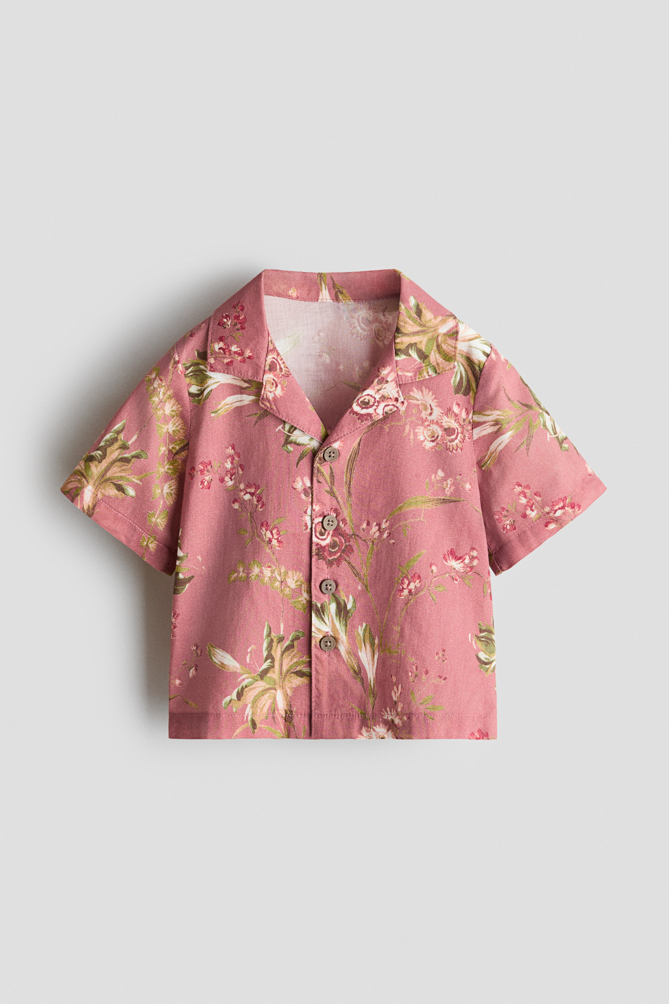 View larger image: A short-sleeve button-down shirt is shown from the front, featuring dusty pink fabric with an all-over green and white botanical floral print, a camp collar, and a front placket with four brown buttons.