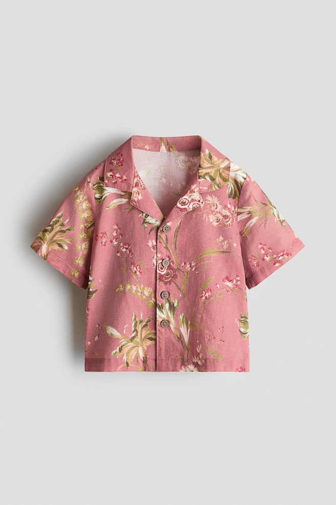 A short-sleeve button-down shirt is shown from the front, featuring dusty pink fabric with an all-over green and white botanical floral print, a camp collar, and a front placket with four brown buttons. - 1