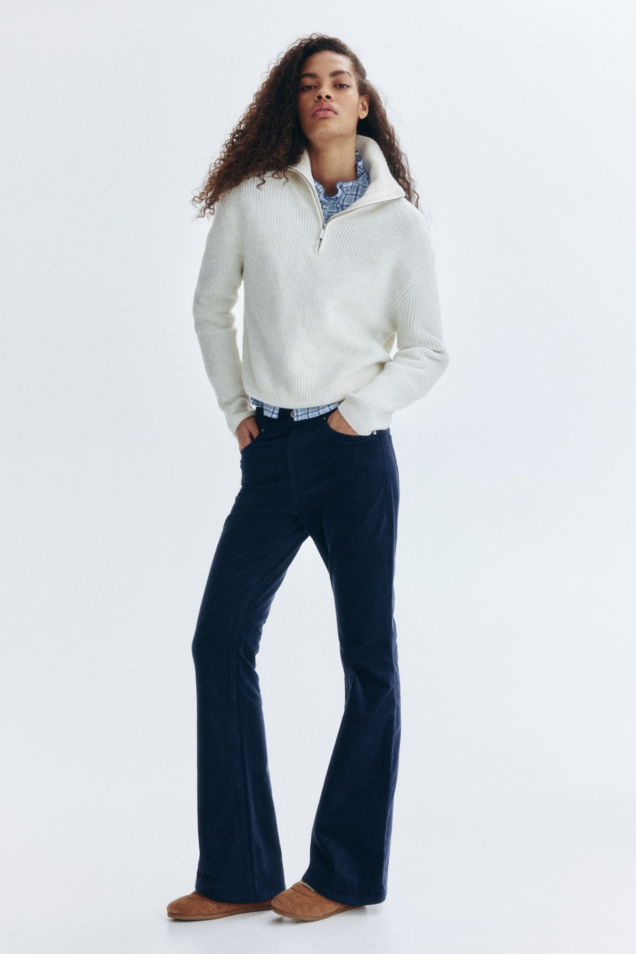 Voir une image plus grande: Un chandail à demi-fermeture éclair en tricot côtelé crème, avec un col montant et une fermeture éclair argentée, est porté par le mannequin. Il est superposé sur une chemise à carreaux bleus, avec un pantalon évasé en velours côtelé bleu marine et des pantoufles marron clair complétant la tenue.