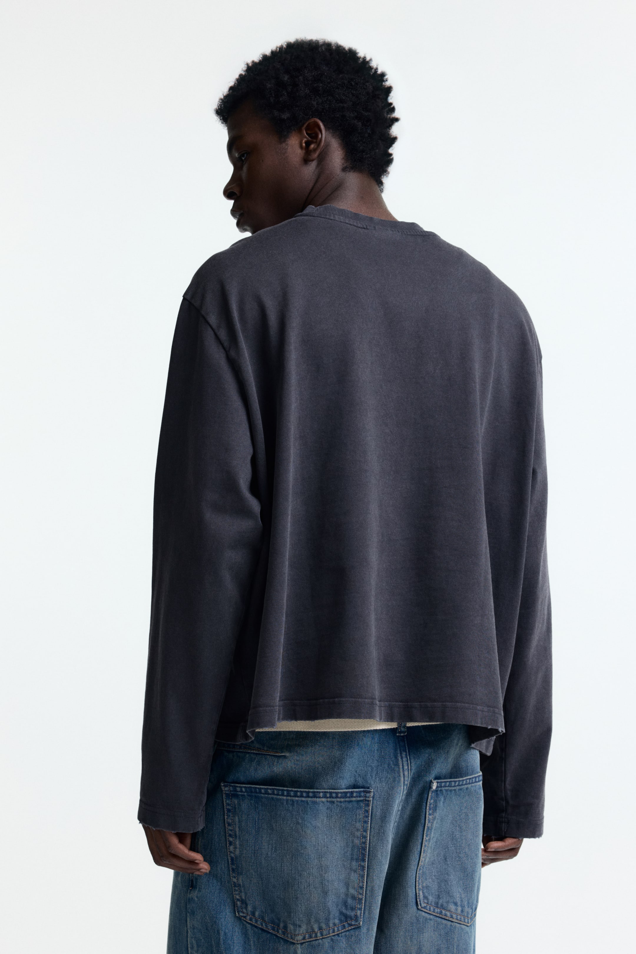 View larger image: A dark gray long-sleeved shirt is being presented from the back, angled slightly to the right, featuring a washed finish and a relaxed fit. It is designed with a crew neck, long sleeves, and a raw, unfinished hem, with a lighter-colored undershirt layering beneath.