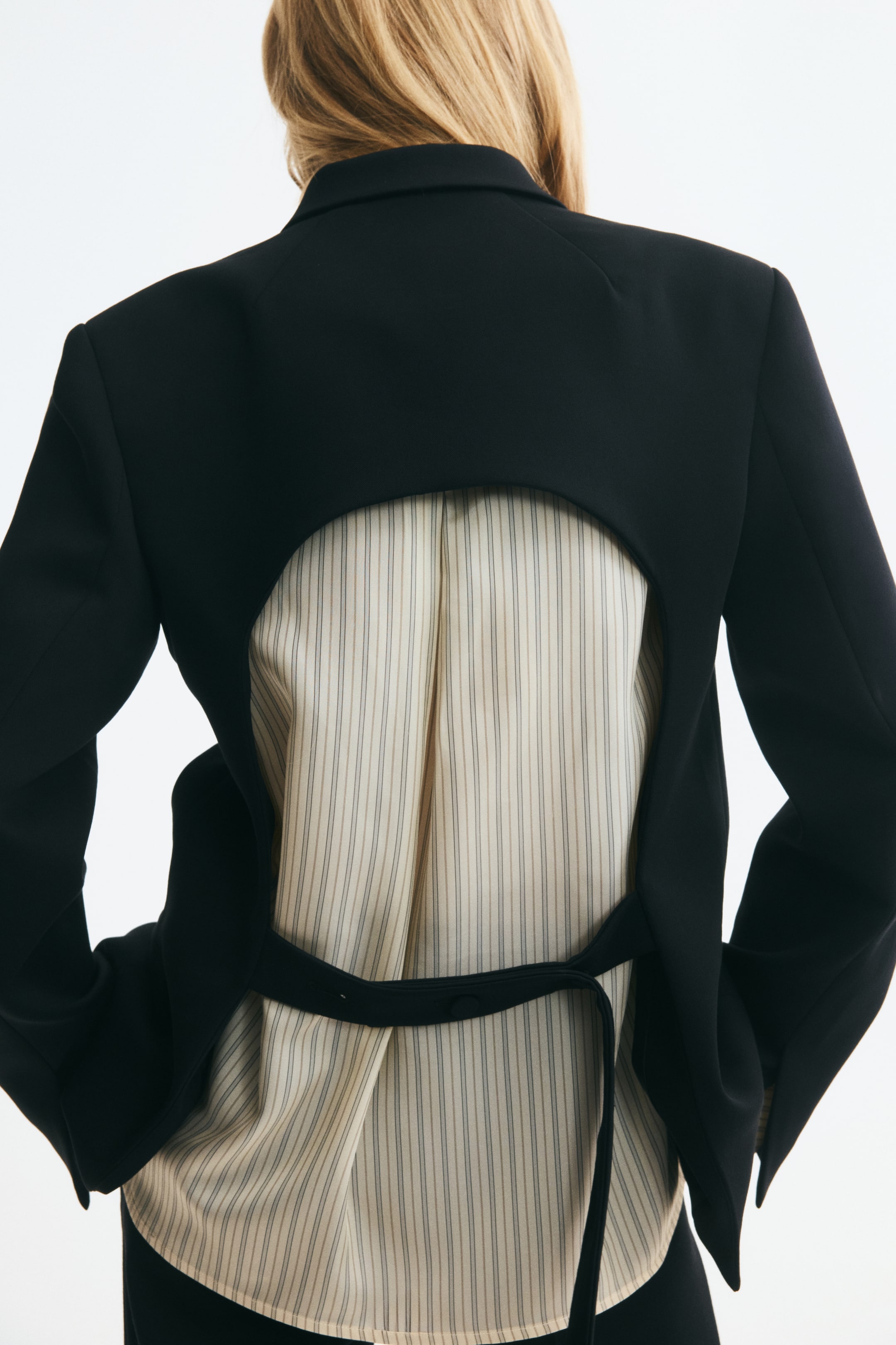 Größeres Bild anzeigen: Ein schwarzer Oversize-Blazer mit teilweise offenem Rücken wird vom Model über einem cremefarbenen, vertikal gestreiften Hemd getragen. Der Rückenausschnitt verfügt über einen horizontalen Riemen, der über den unteren Rücken verläuft.