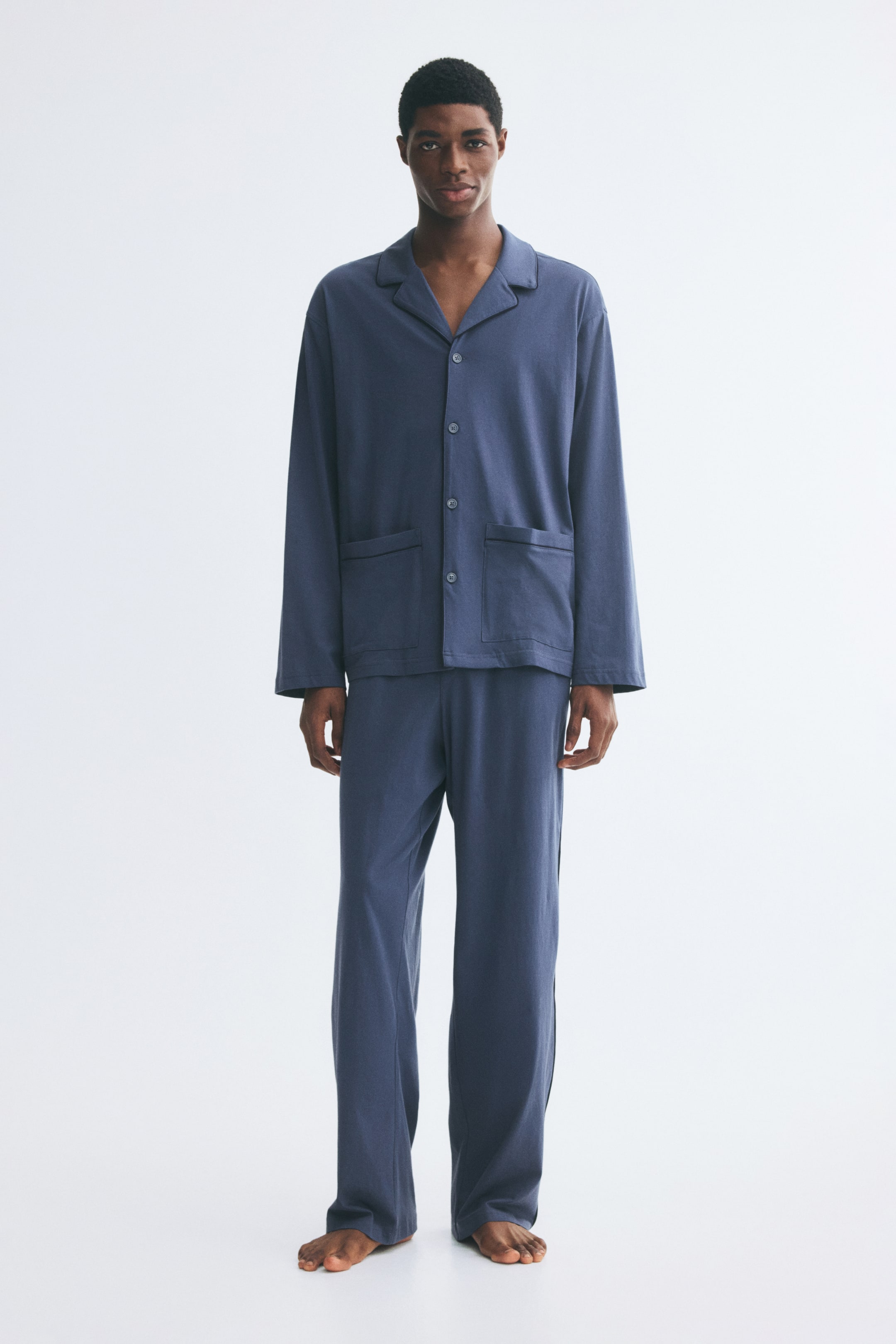 View larger image: The model is wearing a dusty blue pyjama set in soft cotton jersey. The long-sleeve shirt features a resort collar with dark piping, front buttons, and two patch pockets, paired with matching straight-leg trousers.