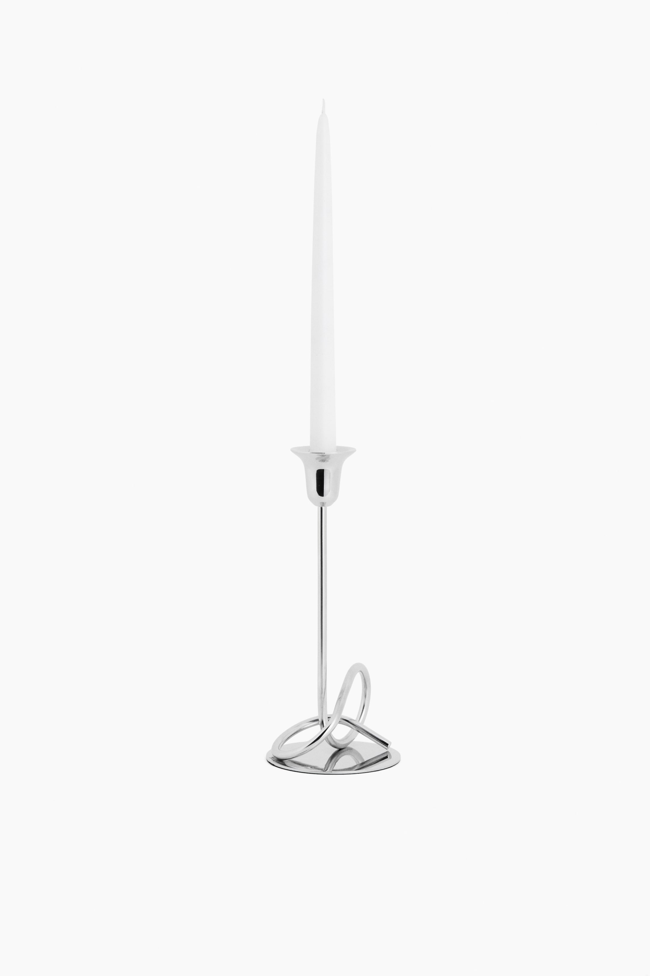 View larger image: A stainless steel candlestick is standing upright from a front-facing angle, featuring a flat, round base and a sculptural, knot-inspired loop design at the bottom of its slender stem. A flared candle holder cup at the top is holding a tall, white taper candle.