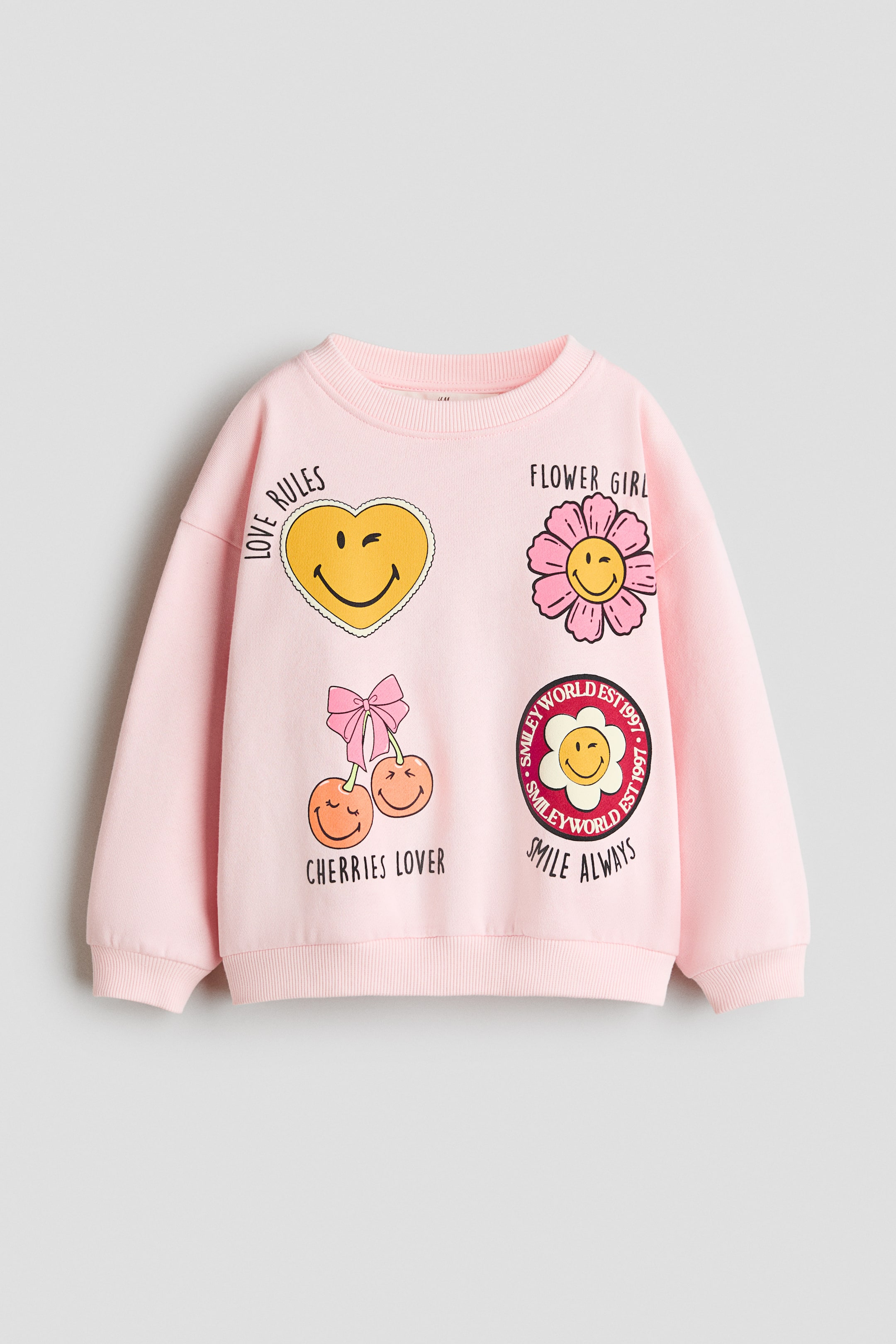Agrandir l'image: Un sweat-shirt rose clair oversize est présenté de face, avec un col rond bordé de côtes, des épaules tombantes et des finitions côtelées aux poignets et à l’ourlet. Le devant affiche quatre motifs imprimés colorés : un smiley cœur jaune, un smiley fleur rose, des cerises souriantes avec un nœud et un badge rond smiley.