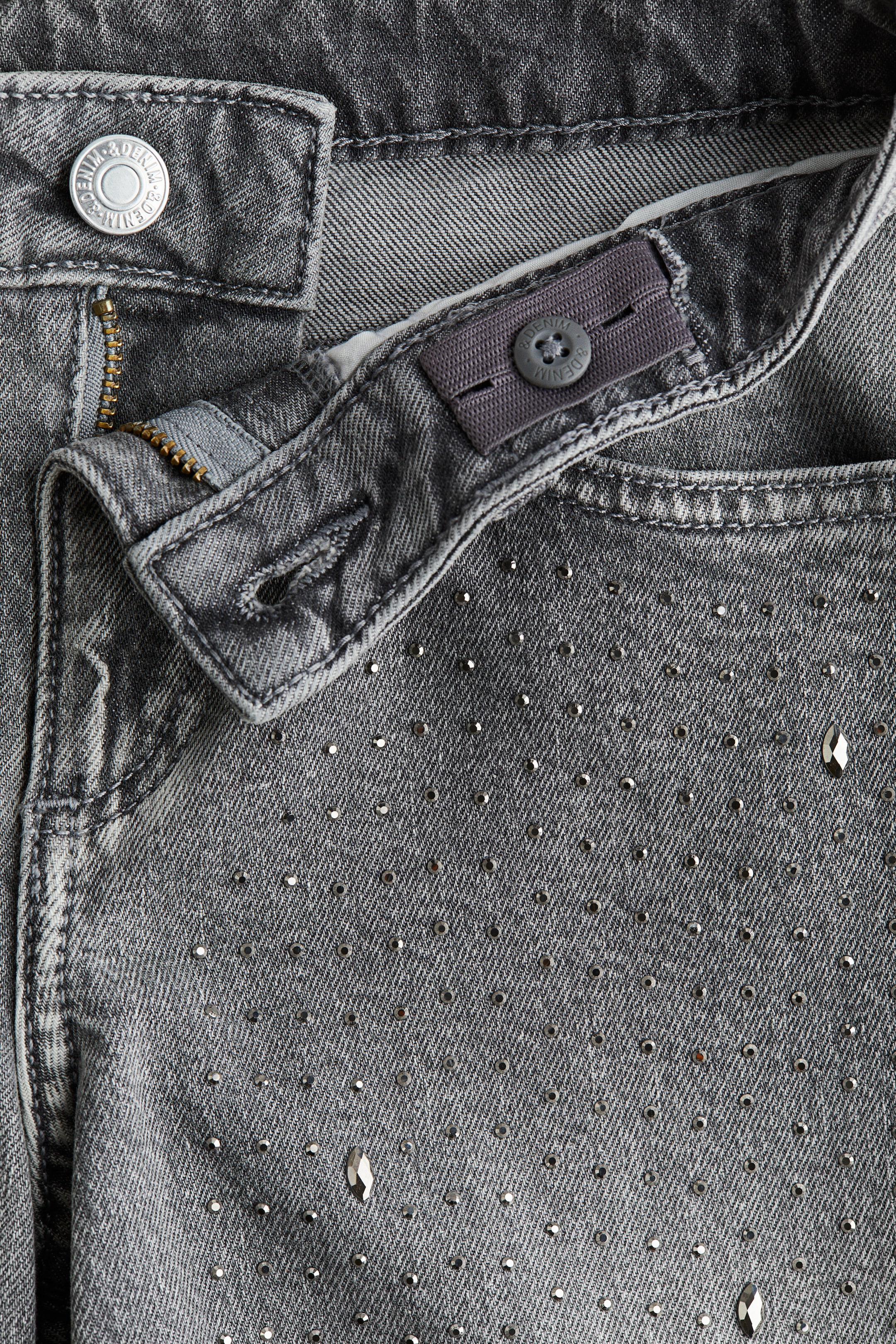 View larger image: Front view of 5-pocket gray cotton denim jeans with a washed-out look, featuring a low waist with a metal button and zip fly closure. An adjustable elastic band with a button is being seen on the inner waistband, and the denim fabric is being embellished with numerous small, round metallic studs and scattered larger, faceted rhinestones across the front.