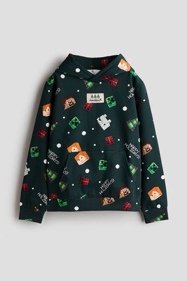 Una felpa con cappuccio verde scuro è mostrata frontalmente, con una stampa natalizia all-over con personaggi pixelati di Minecraft, alberi di Natale e la scritta "HAPPY HOLIDAYS!". La felpa presenta un cappuccio avvolgente, spalle scese, una tasca a marsupio frontale e polsini e orlo a coste. - 1