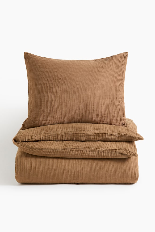 A brown cotton muslin quilt cover set is shown from the front, featuring a matching pillowcase standing upright behind a neatly folded quilt cover. Both pieces display a distinct crinkled, puckered texture. - 1