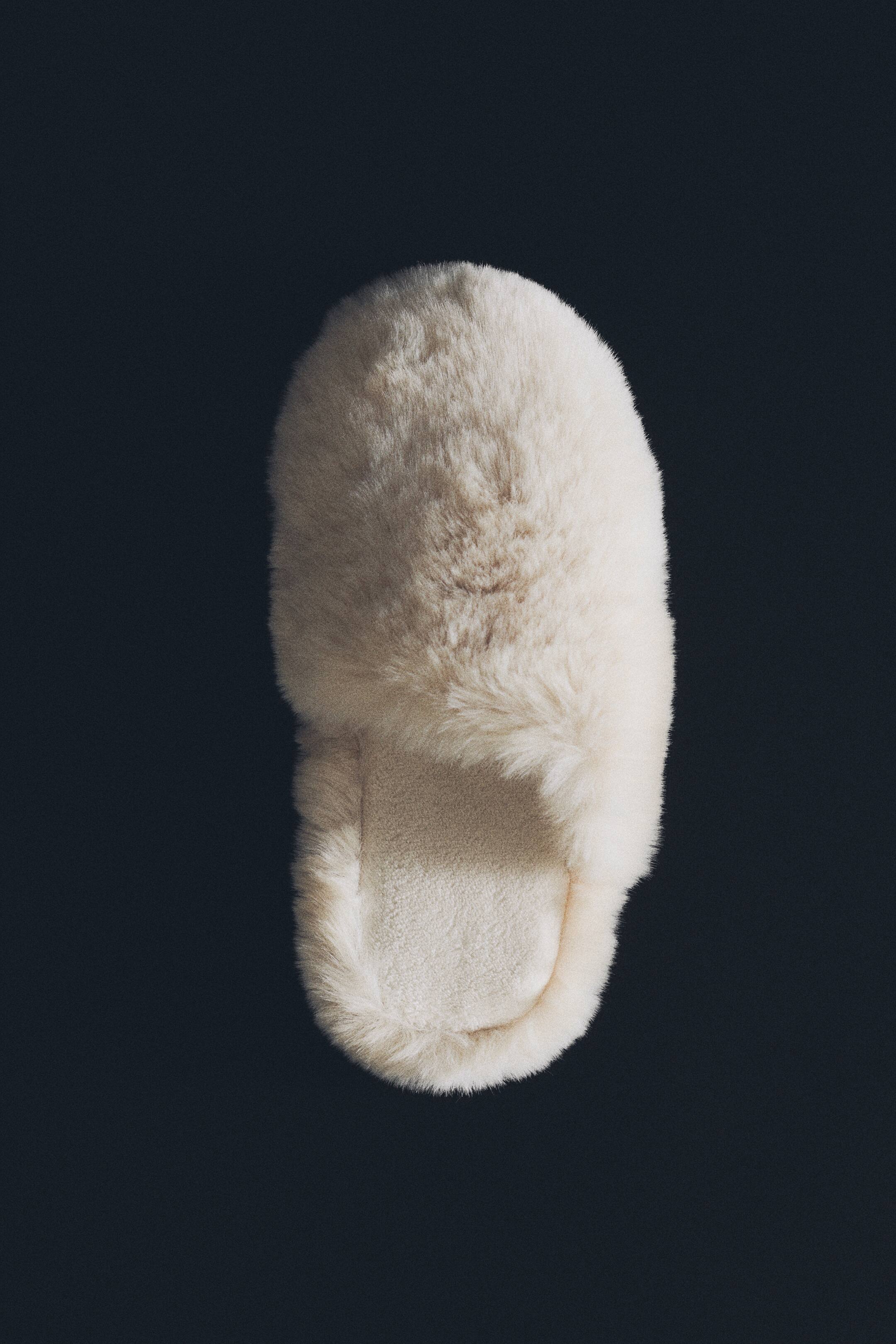 View larger image: A single off-white slipper in a soft, fluffy fabric is shown from a top-down view. The slipper features a rounded closed toe, a plush faux-fur upper, and a smooth, velour-lined footbed.