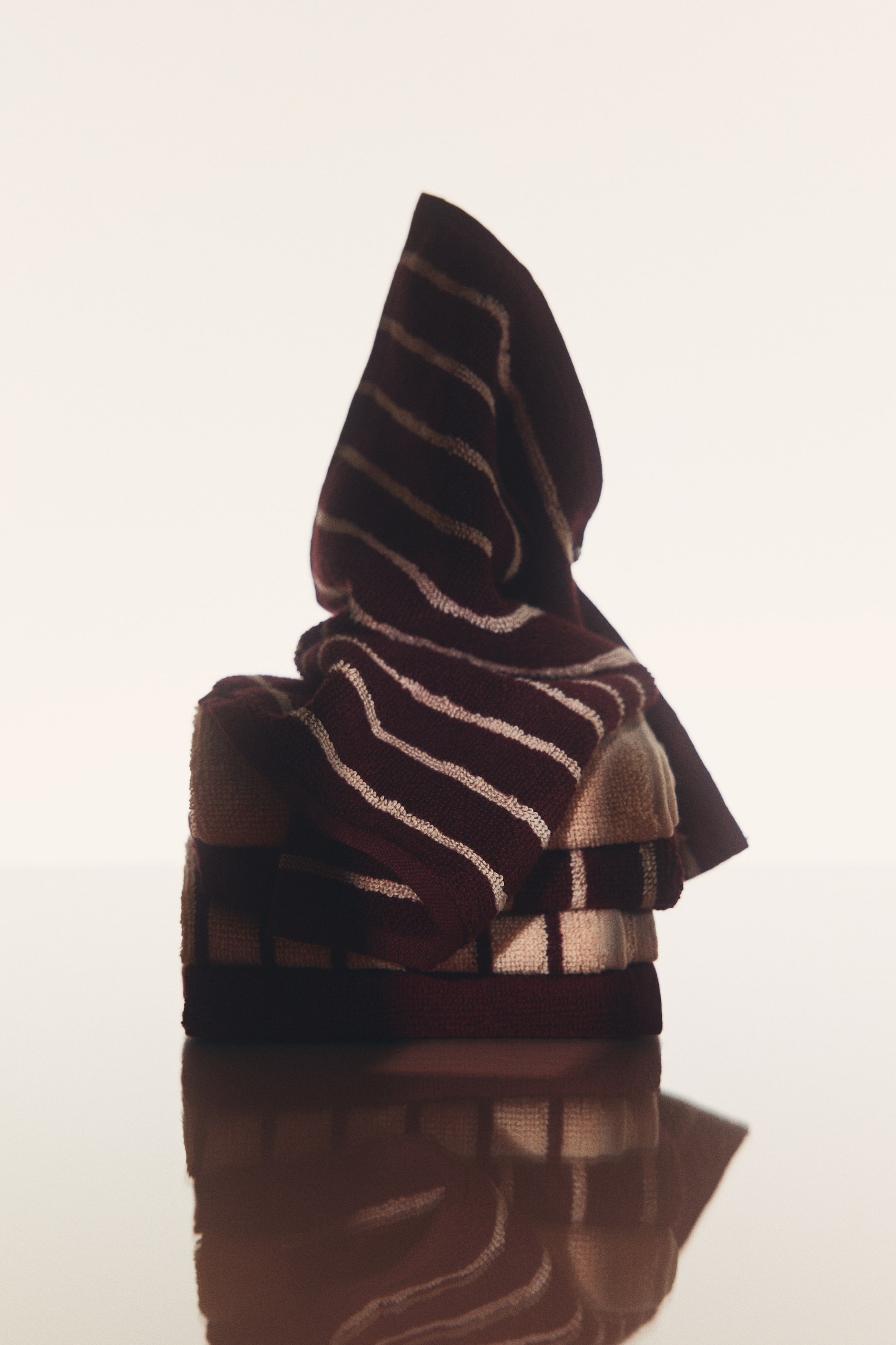 View larger image: A set of cotton terry face cloths in solid burgundy, cream, and striped designs is stacked on a reflective surface. One striped face cloth is draped upright over the top of the stack, showcasing its soft, textured fabric from a front angle.