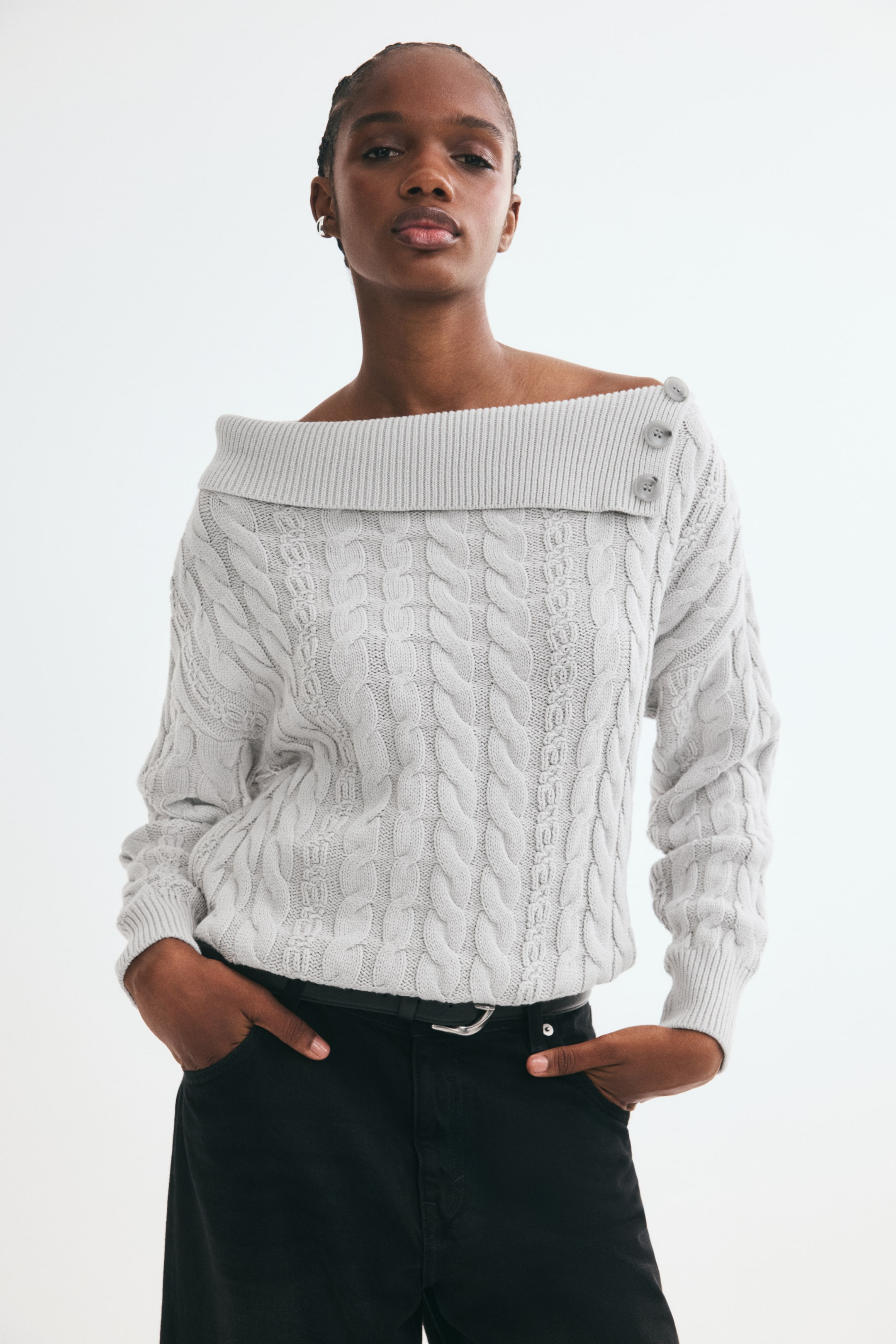 Xem ảnh lớn hơn: Người mẫu đang mặc một chiếc áo len dệt cáp màu xám nhạt dáng oversized, nổi bật với phần cổ gập lệch bất đối xứng có gân và ba khuy trang trí trên một bên vai. Áo được phối sơ vin vào quần đen có thắt lưng, làm nổi bật phom dáng thoải mái cùng phần bo tay áo có gân.