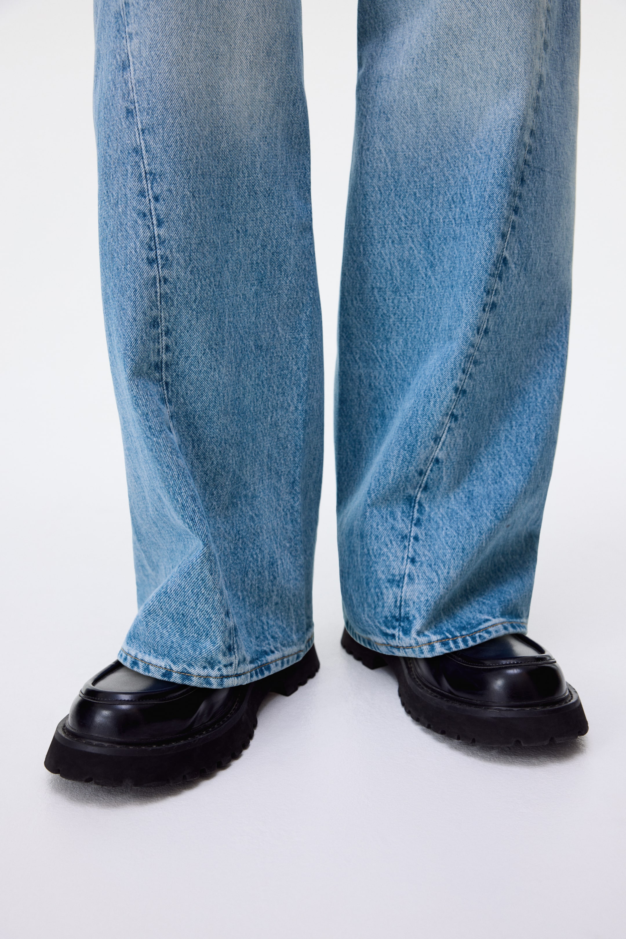 View larger image: Light blue, wide-leg denim jeans are draping over black chunky-soled loafers with moccasin stitching, all facing forward.