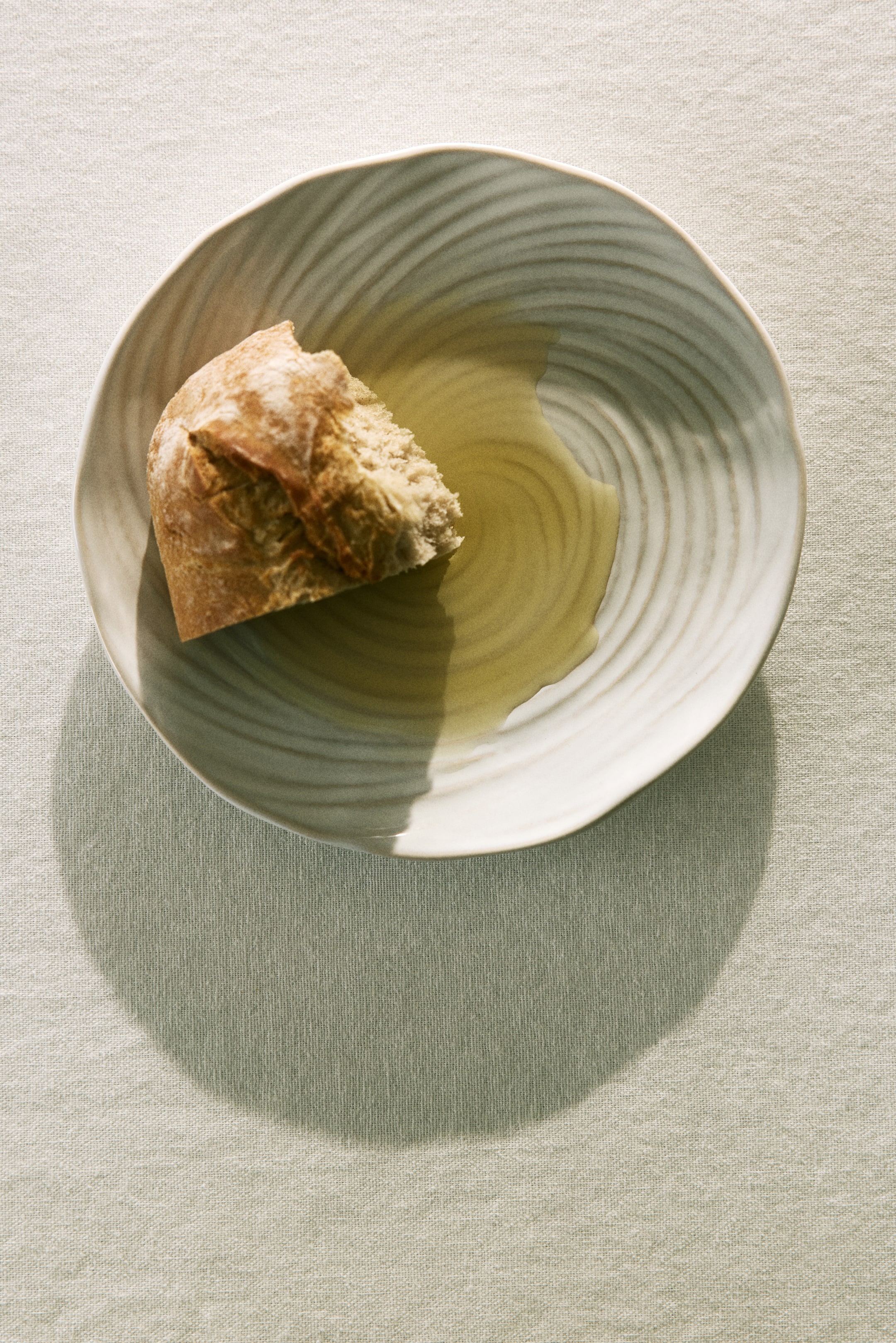 View larger image: A piece of rustic bread is resting in a shallow, light-coloured dish with subtle concentric ridges, partially submerged in golden olive oil.