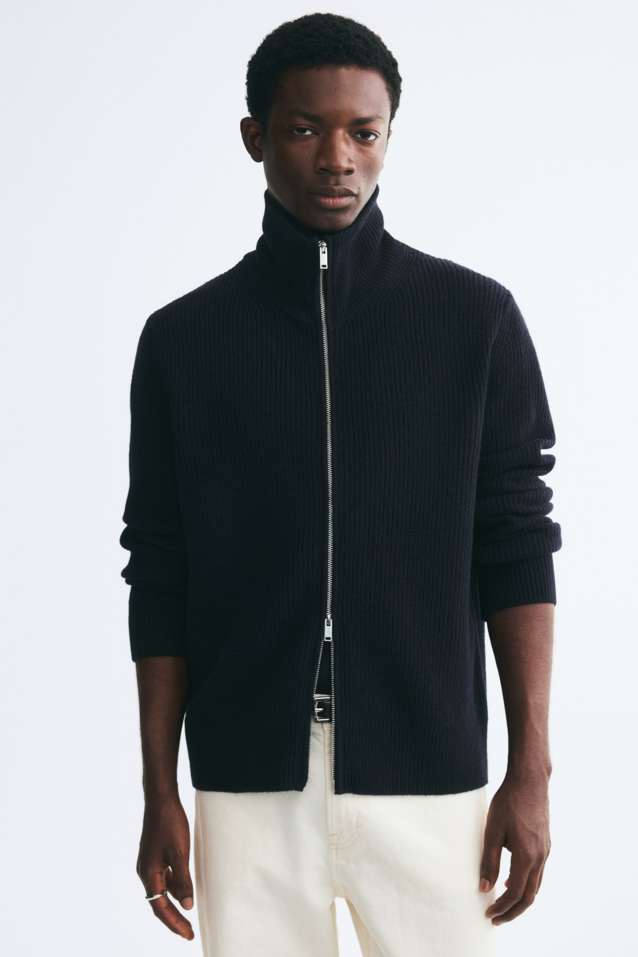 View larger image: The model is wearing a dark navy rib-knit cardigan featuring a stand collar, long sleeves, and a straight hem. The cardigan is styled with its two-way silver zipper closed up to the neck and slightly unzipped at the bottom, paired with cream-colored trousers.