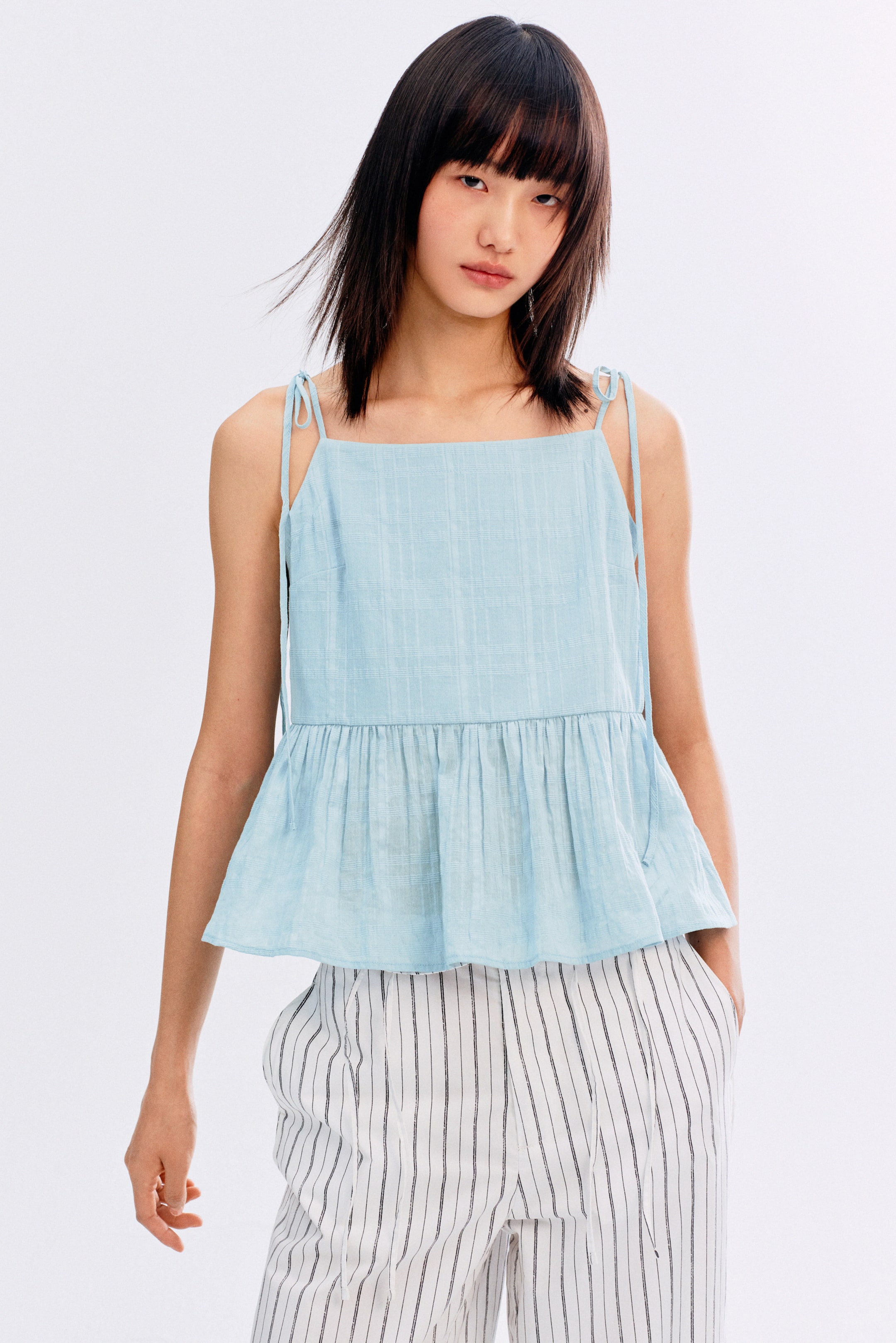 View larger image: A light blue peplum top na may square neckline, manipis na tie straps, at gathered flounce hem, na may subtle grid texture ang tela. Ito ay suot sa ibabaw ng puting pantalon na may manipis na vertical na itim na guhit at drawstring waist.