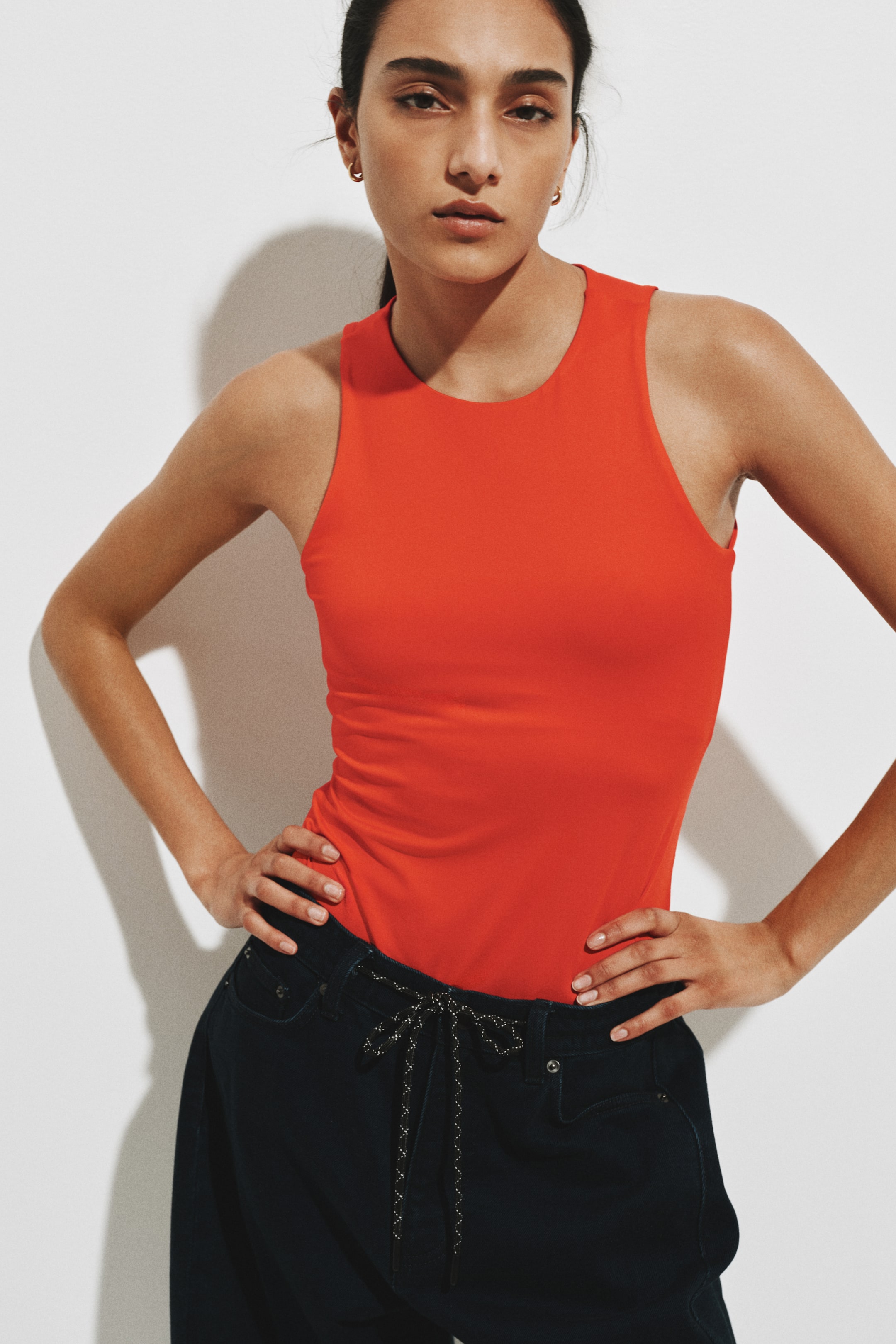 View larger image: A vibrant red sleeveless top is being worn, featuring a round neckline, wide shoulder straps, and a form-fitting silhouette, tucked into dark pants.