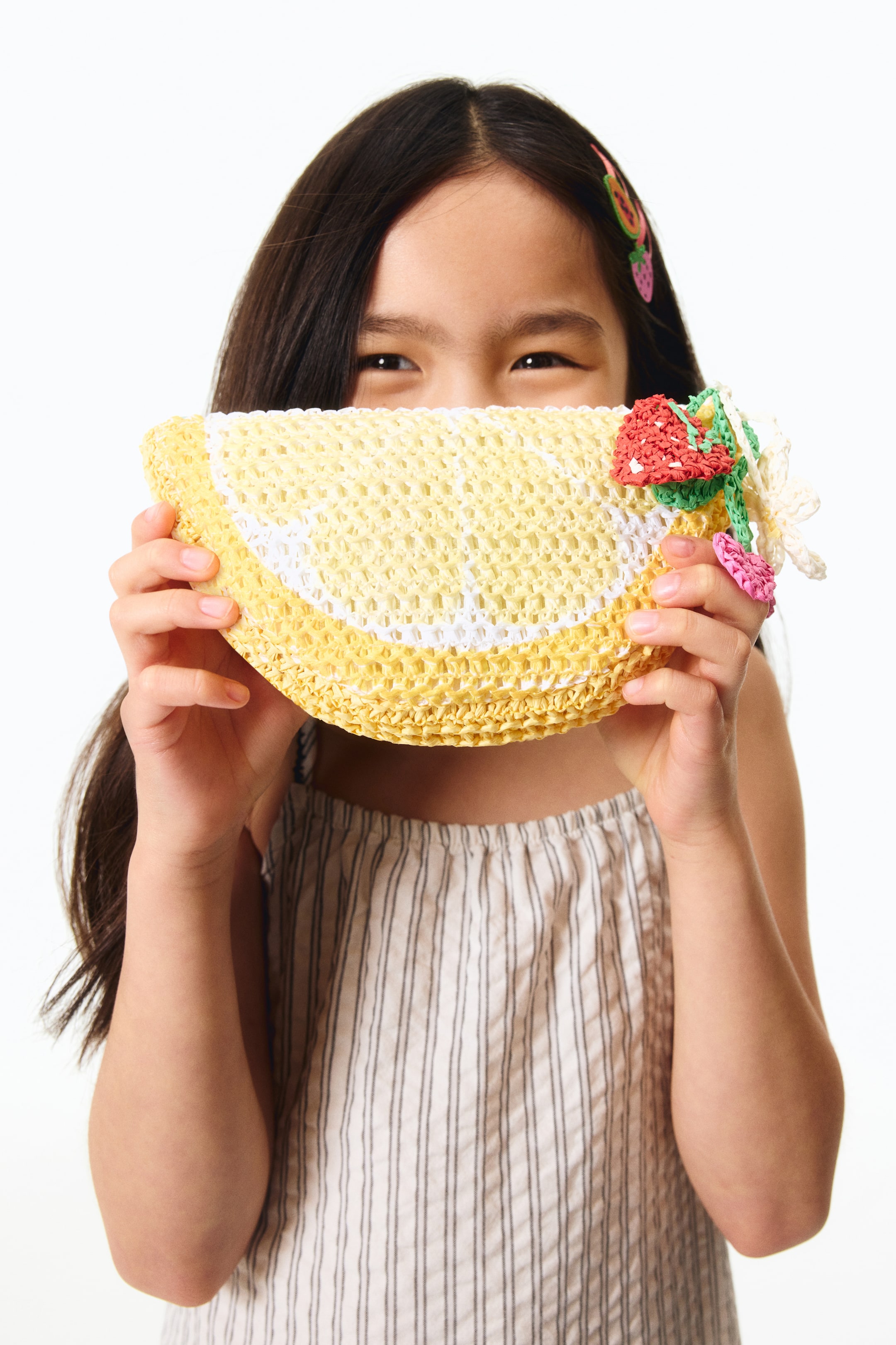 View larger image: A lemon-slice-shaped woven pouch is being held horizontally by a pair of hands. It features a yellow and white crocheted design that mimics a lemon, and a small colorful woven charm with red, green, and pink details is attached to one end.