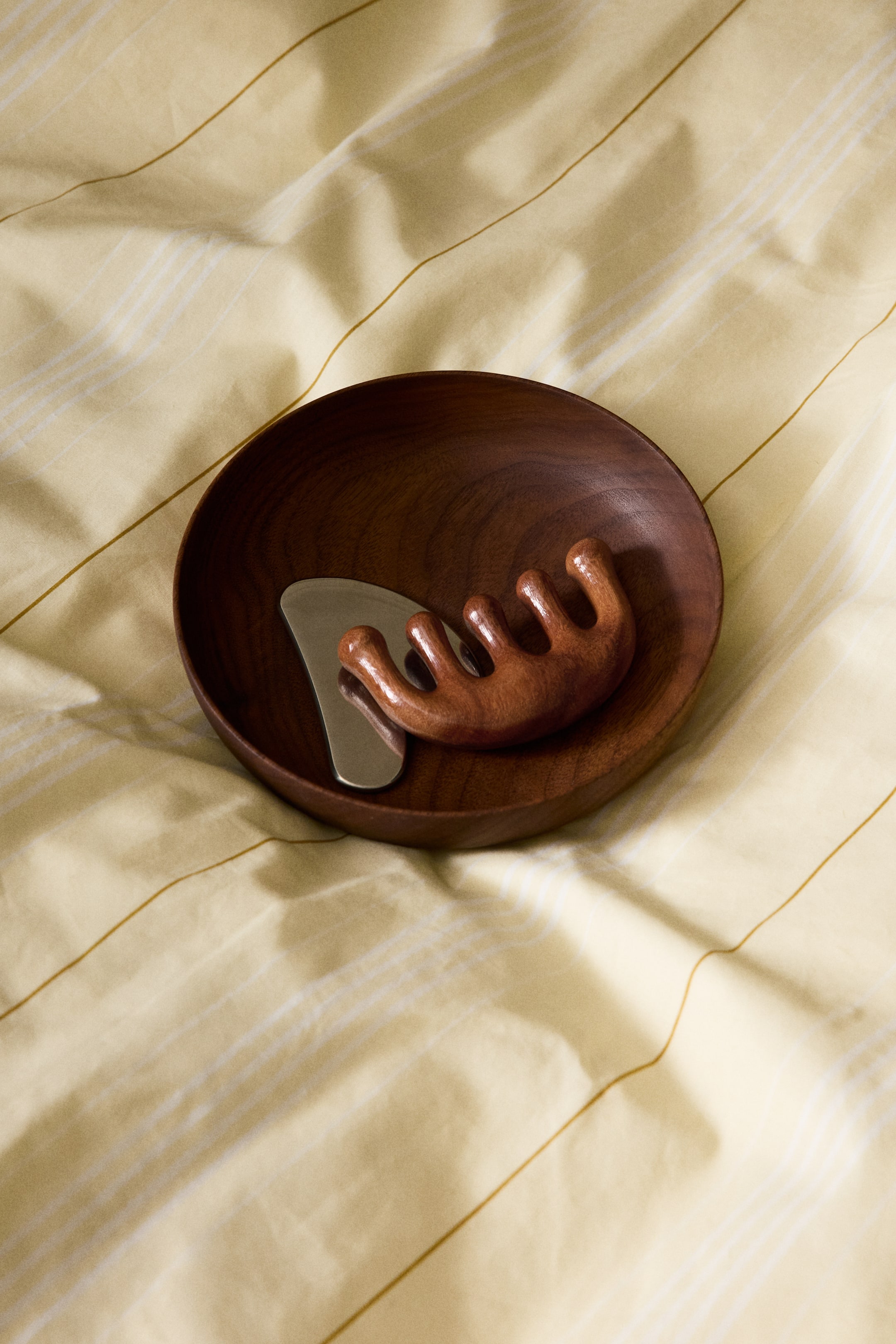 View larger image: A dark brown wooden bowl showcases a silver-toned metal gua sha tool alongside a wooden scalp massager. The uniquely shaped metal gua sha tool lies flat, while the five-pronged wooden scalp massager rests on its side within the bowl.