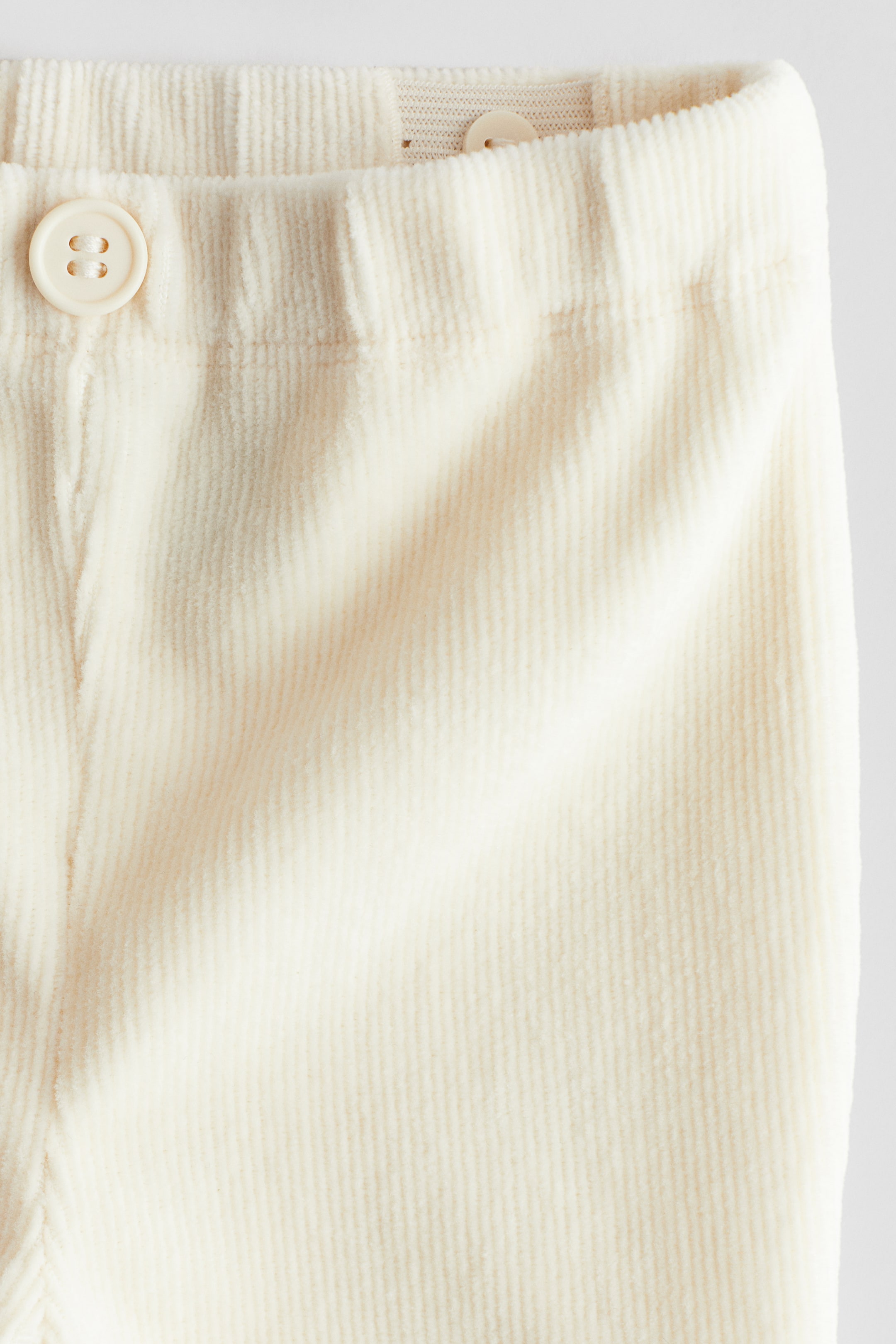 View larger image: The front of cream corduroy pants is displaying prominent vertical wales. A subtly gathered waistband is visible at the top, featuring a cream four-hole button on the left and a smaller cream button partially visible on the upper right.
