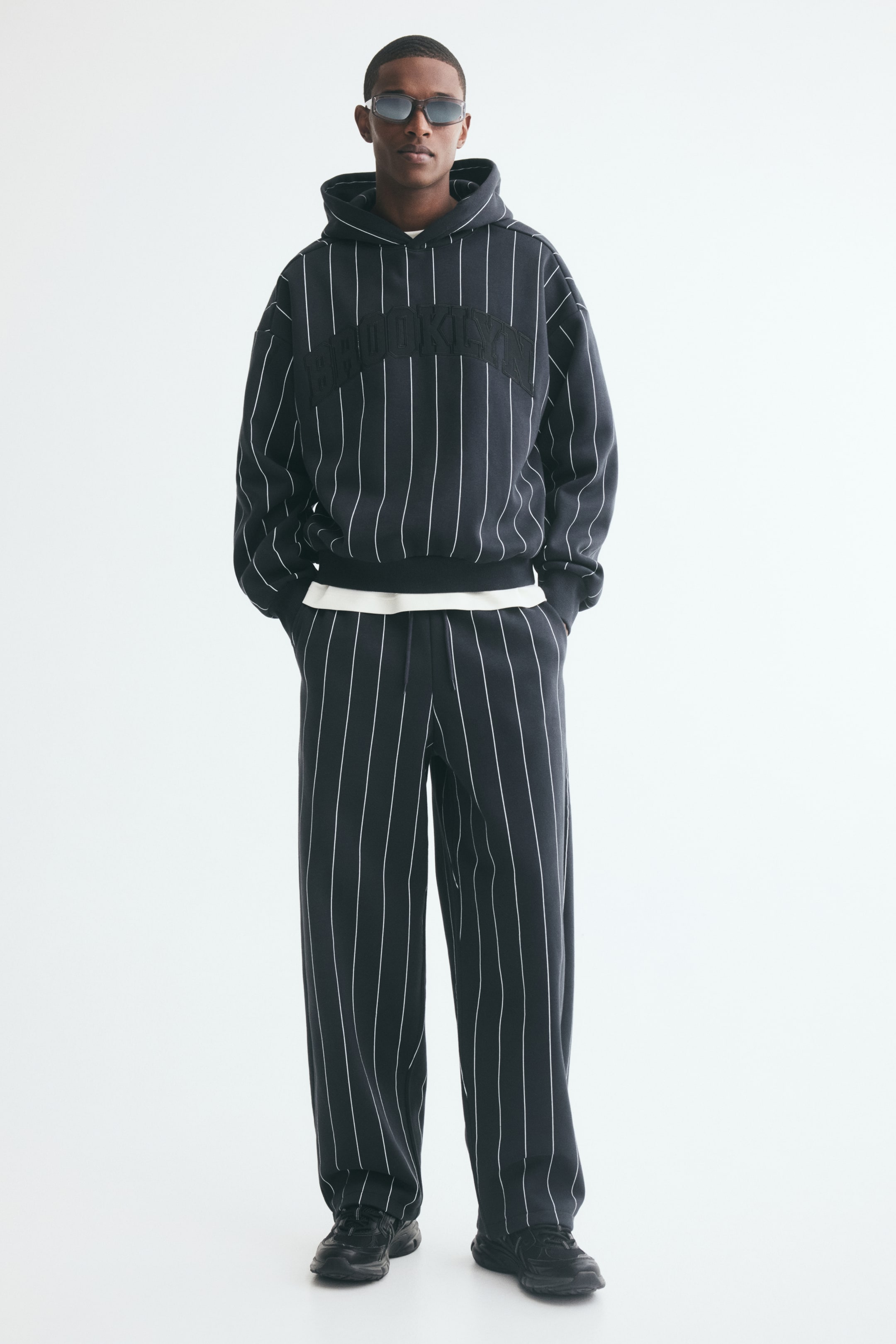 View larger image: Pinstriped track pants in dark grey fleece fabric are worn by the model with hands placed in the diagonal side pockets. The pants feature a relaxed fit, barrel legs, and an elasticated waist, and are styled with a matching pinstriped hoodie and black sneakers.