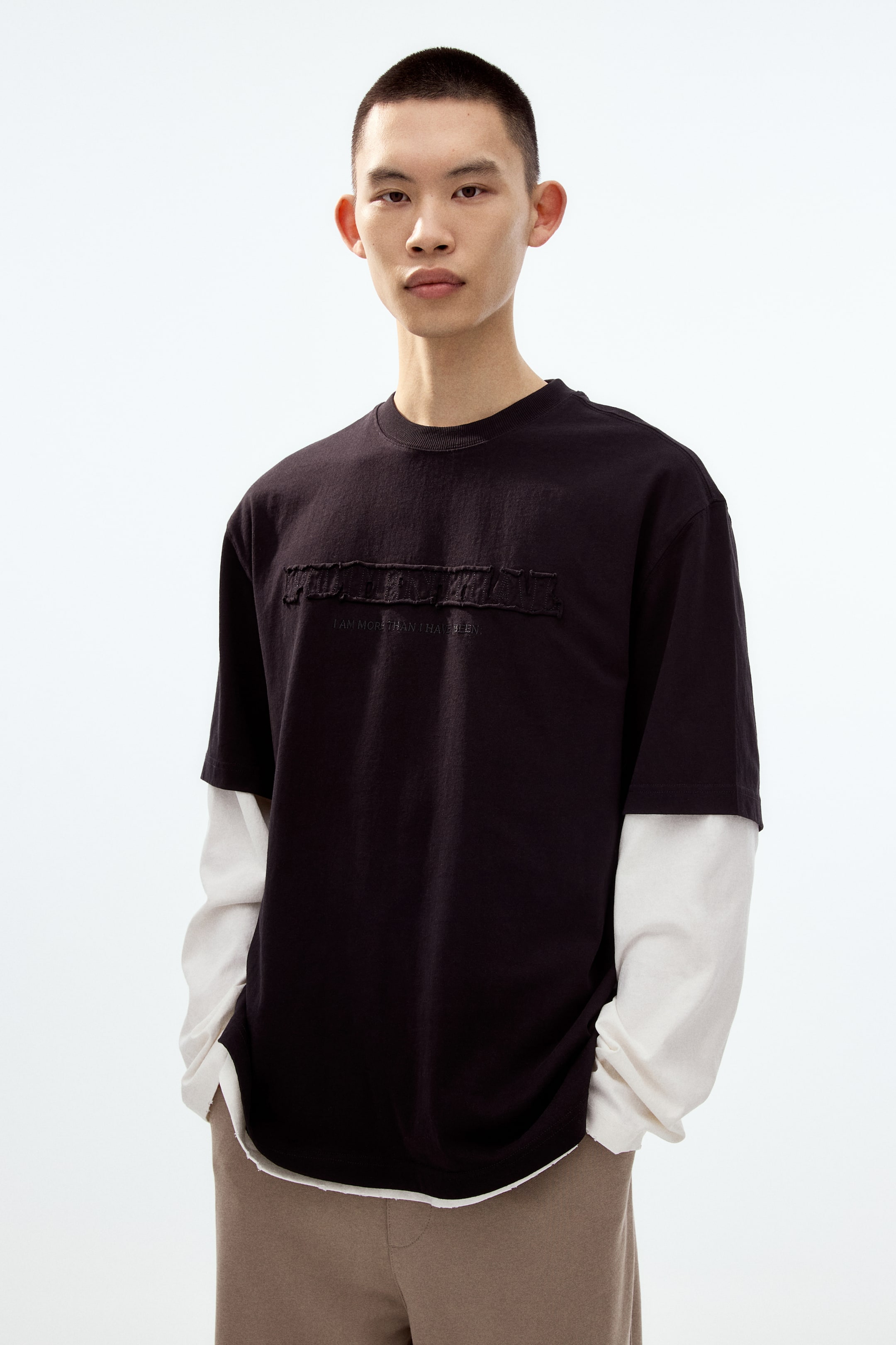 View larger image: A black short-sleeved t-shirt features a ribbed crew neck and a textured, distressed horizontal appliqué on the chest, with "I AM MORE THAN I HAVE BEEN" embroidered beneath it. It is layered over an off-white long-sleeved t-shirt, visible at the short sleeves and below the raw hem of the black t-shirt, shown from a slightly angled front view.