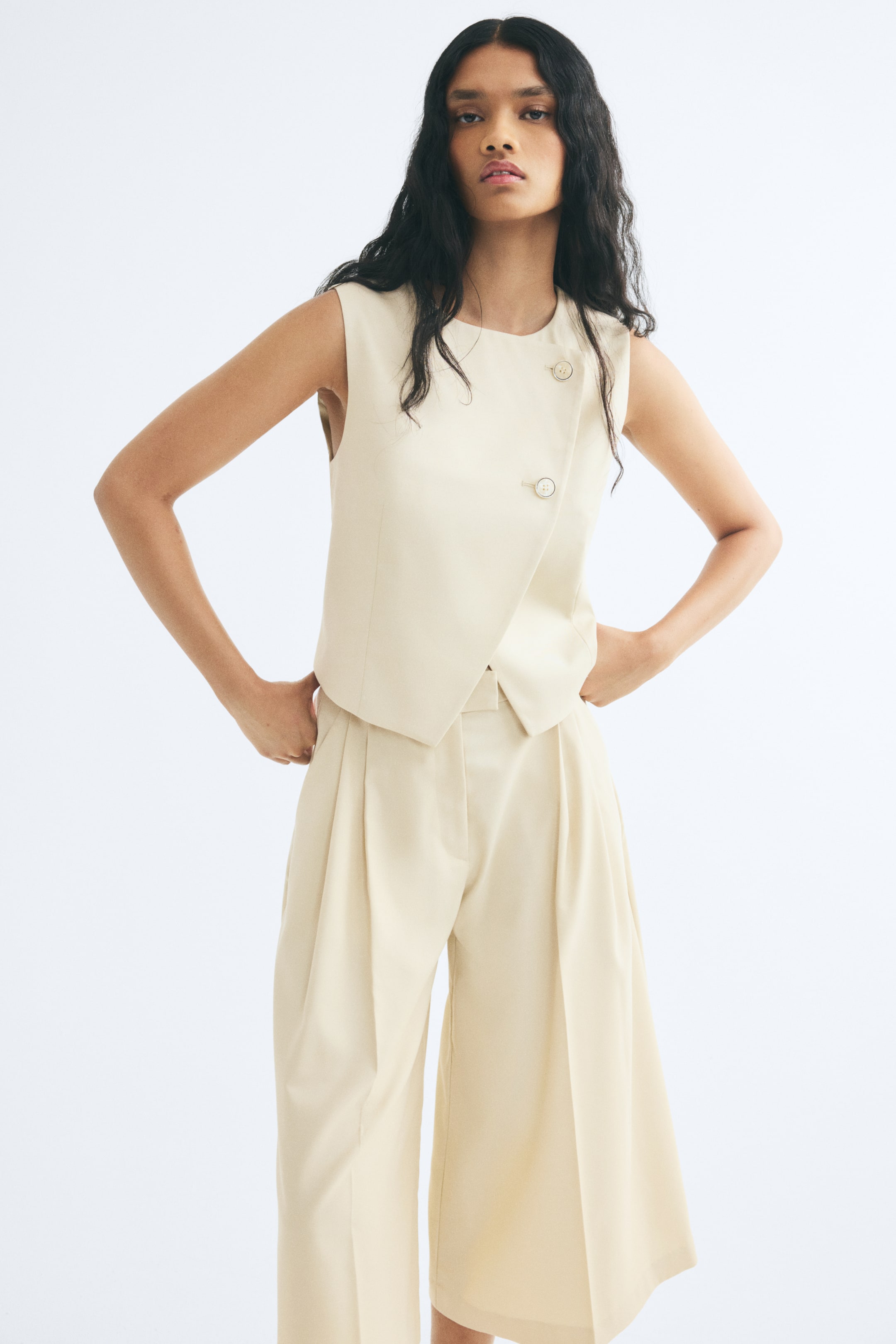 Xem ảnh lớn hơn: Mẫu đang mặc quần culottes dáng rộng màu be nhạt với cạp cao, ly trước và ống rộng có ly là phẳng. Quần culottes được phối cùng áo vest không tay màu kem đồng bộ khi người mẫu đứng với hai tay chống hông.