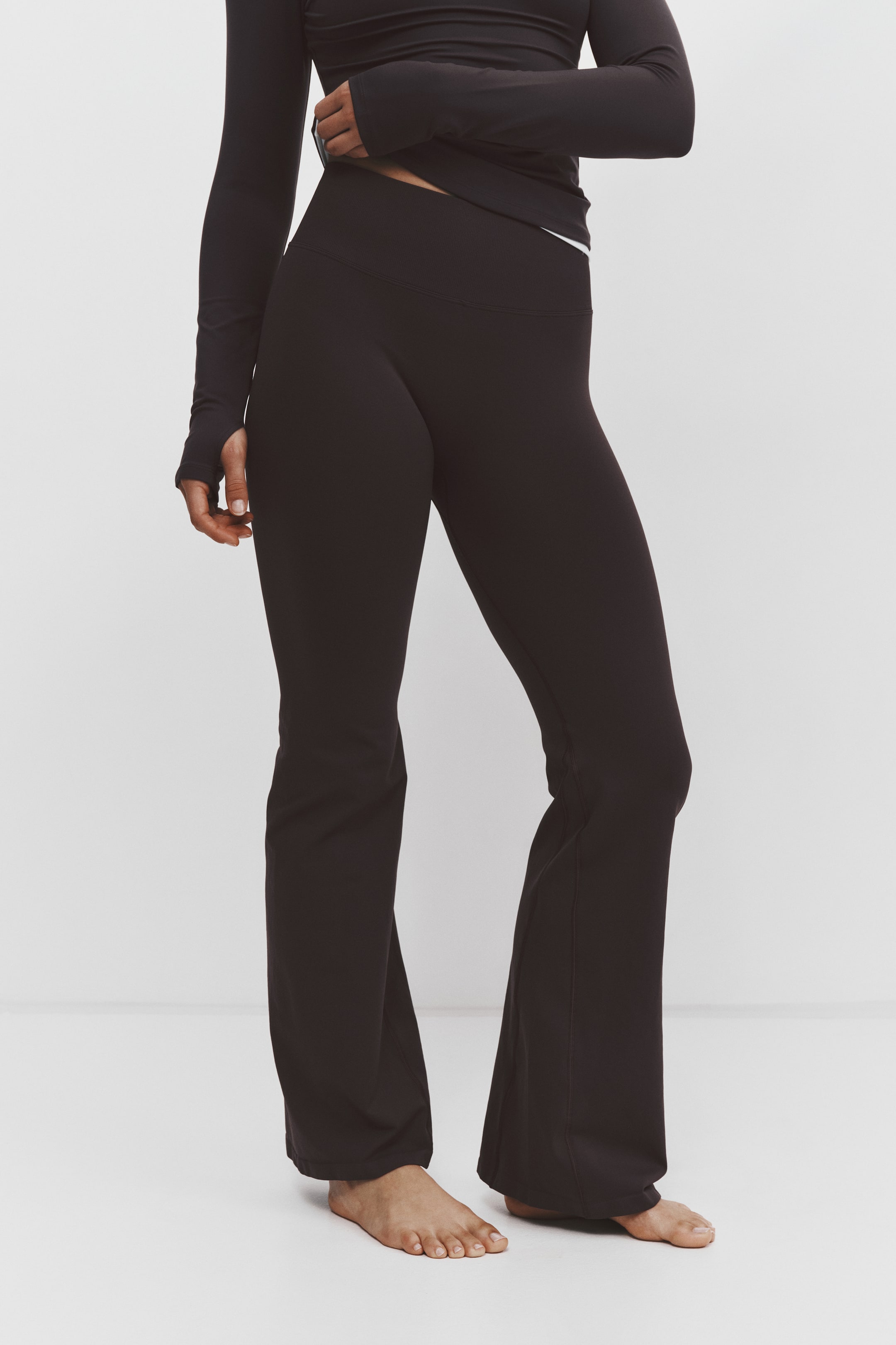 View larger image: Black flared leggings are shown from the front, featuring a high-rise ribbed waistband and a fitted leg silhouette that flares outward from the knee. The leggings are styled with a matching black long-sleeve top that is slightly pulled up to showcase the waist design.
