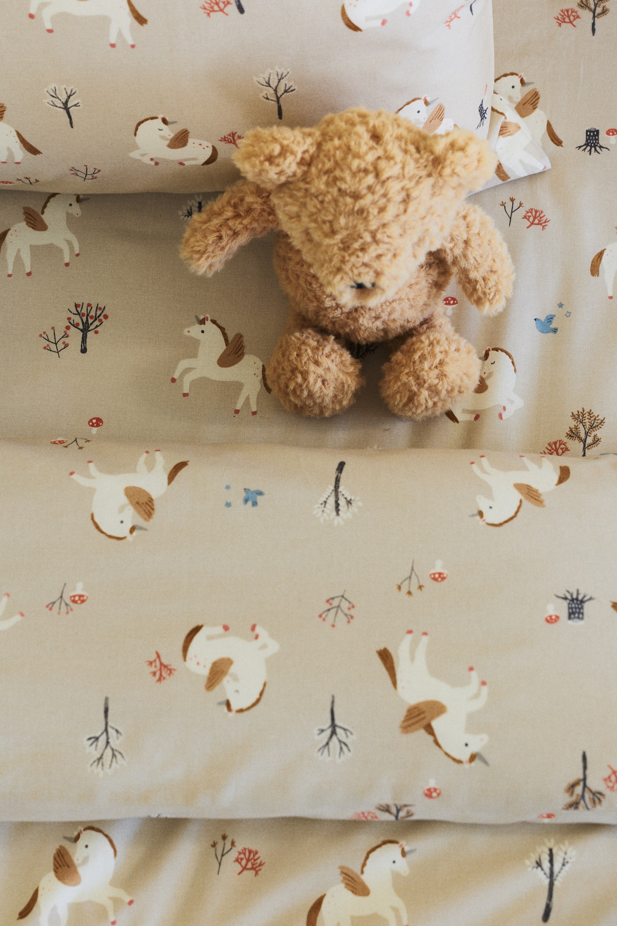 View larger image: Light beige bed linens feature a repeating pattern of white and brown winged horses, small trees, and mushrooms. A fluffy, light brown teddy bear rests on the folded linens.