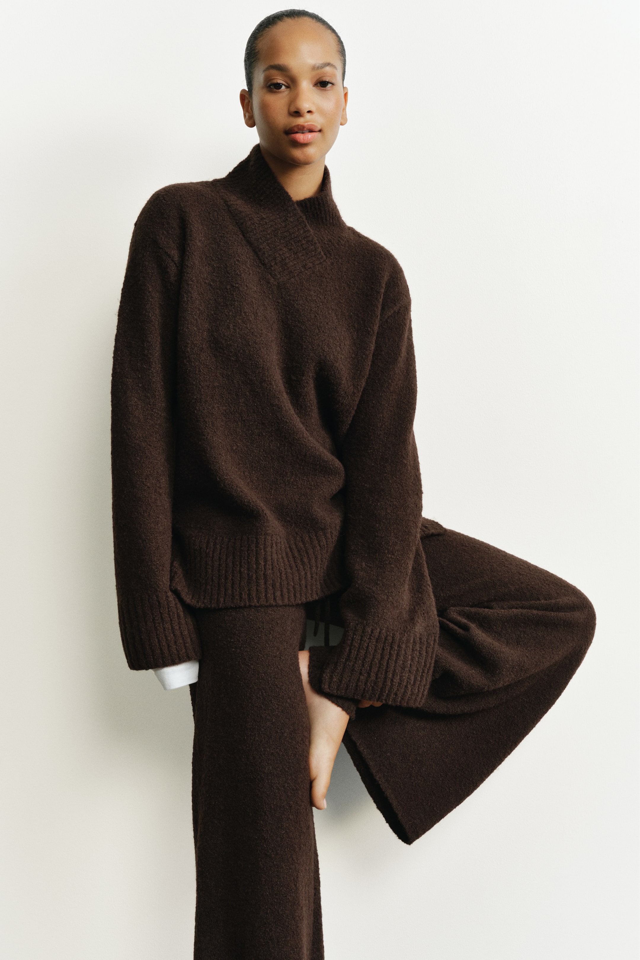 View larger image: A dark brown knit set, consisting of a textured funnel-neck sweater and matching wide-leg trousers, is worn by the model. The sweater features an overlapping high collar and ribbed cuffs, with a white cuff peeking out from one sleeve.