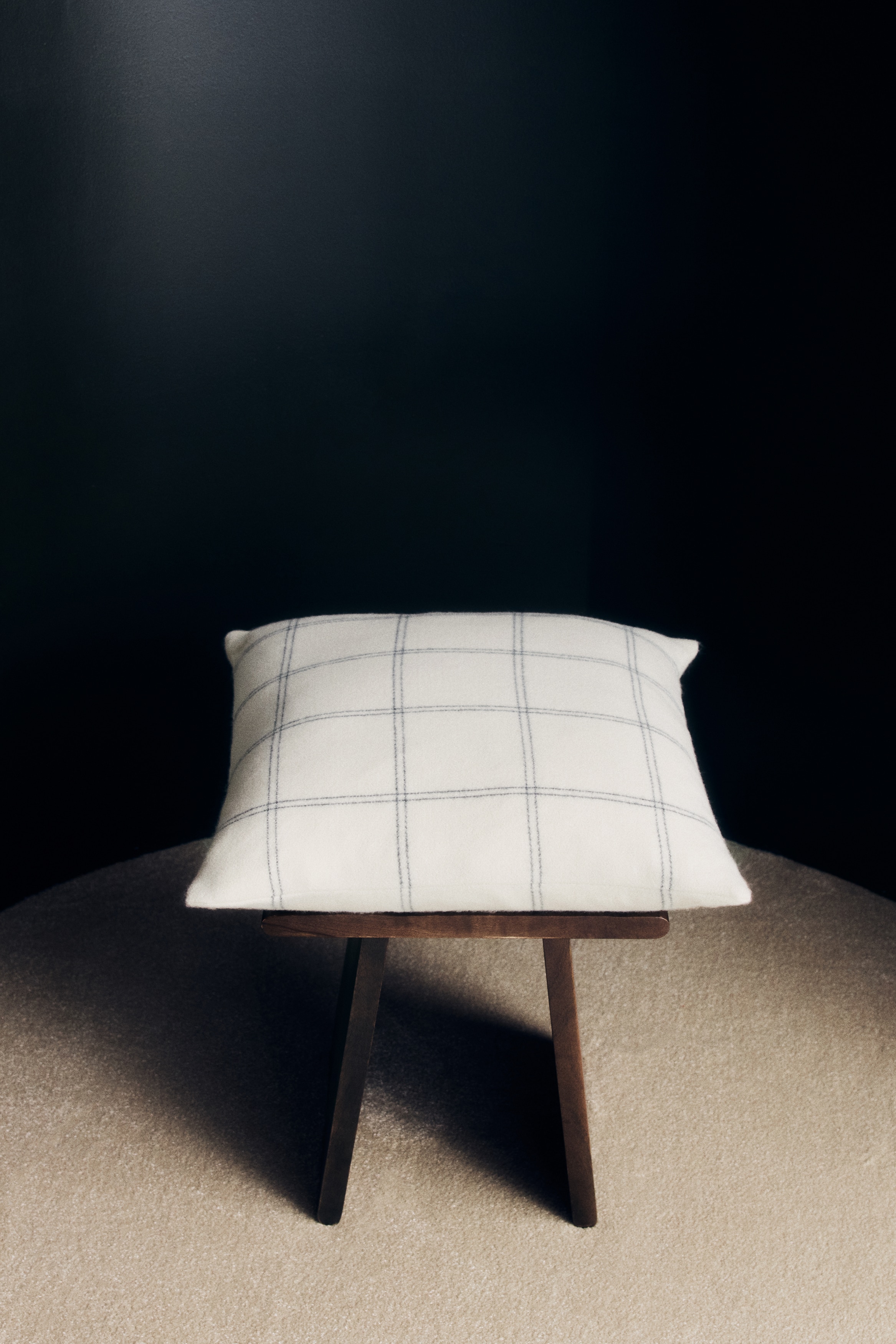 The square cushion is featuring an off-white fabric and a dark, slightly irregular windowpane plaid pattern.