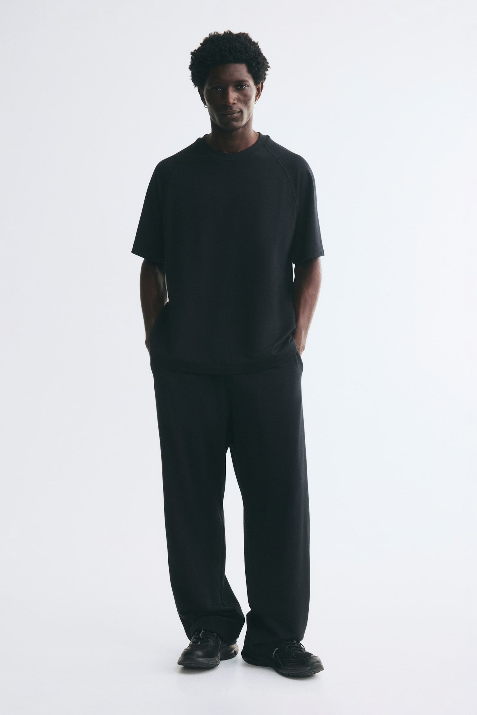 Black track pants featuring a loose-fit silhouette are being worn by the model, who is standing with hands placed in the side pockets. The pants are styled with a matching black short-sleeved t-shirt and black sneakers. - 1
