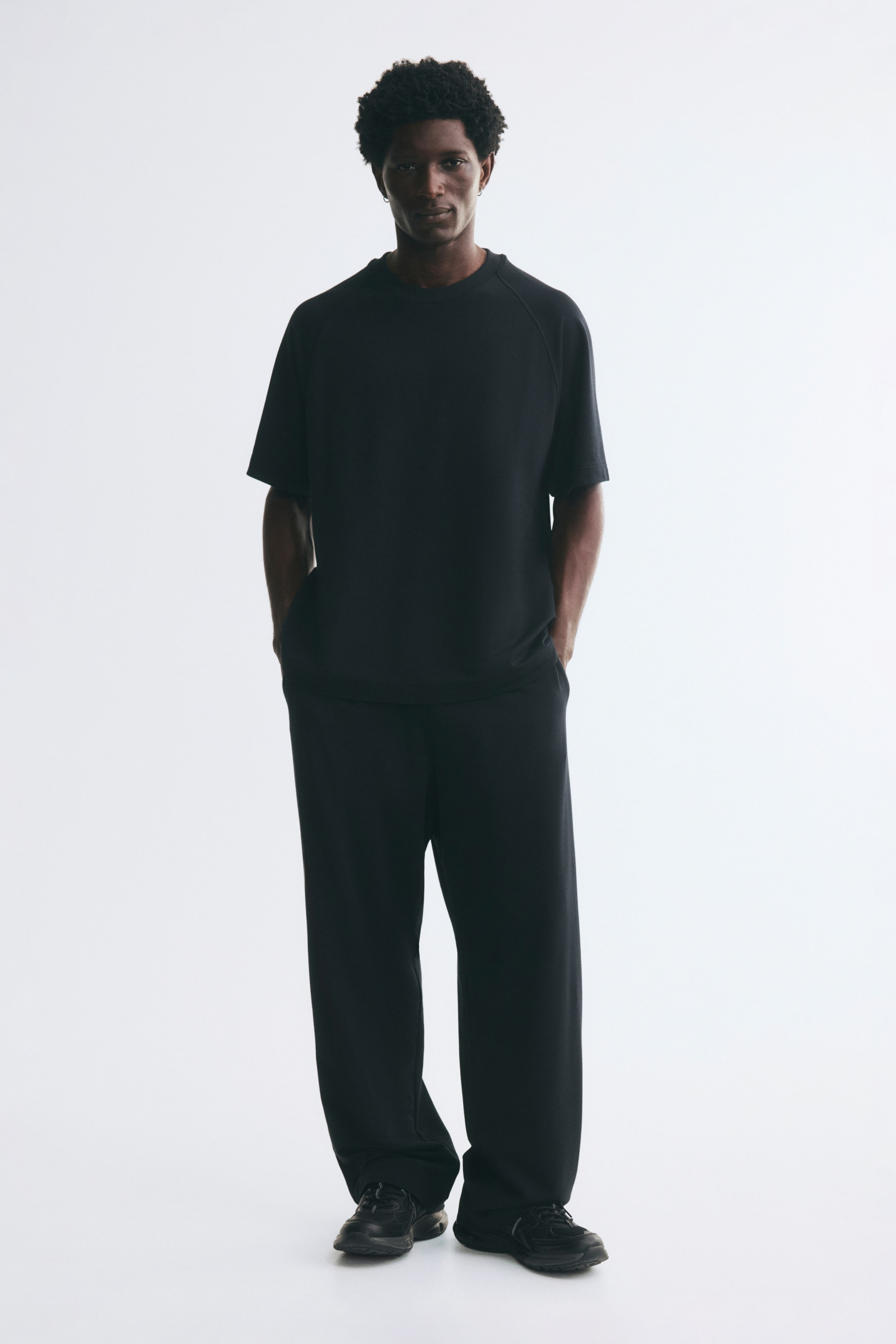 View larger image: Black track pants with a loose-fit silhouette are worn by the model, who is standing with their hands in the side pockets. The pants are styled with a matching black short-sleeved T-shirt and black sneakers.