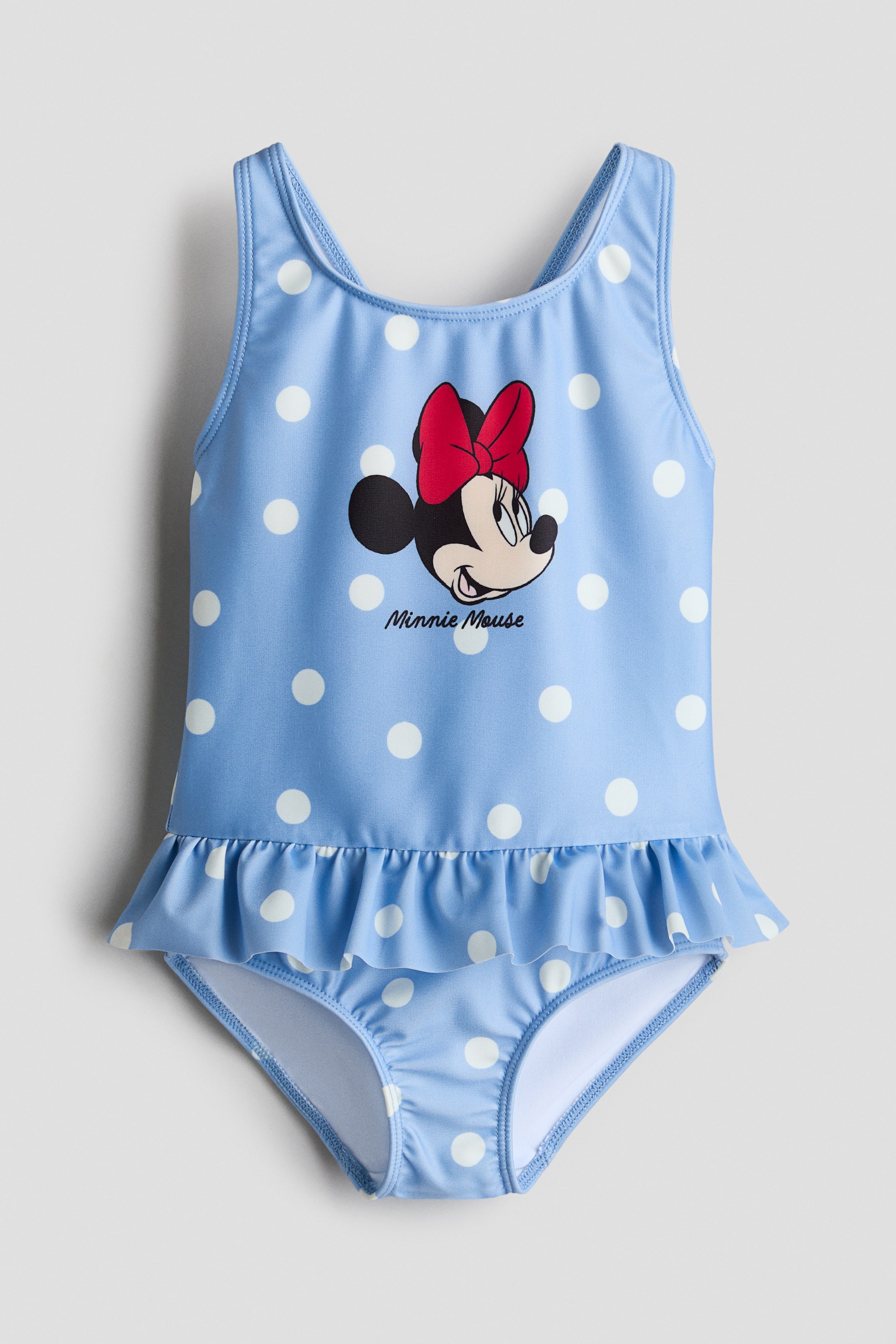 View larger image: The printed swimsuit is displayed front-on, featuring a light blue base adorned with white polka dots. It showcases broad shoulder straps, a graphic of Minnie Mouse's head with a signature red bow, the text "Minnie Mouse" beneath it, and a gathered flounce at the waist.
