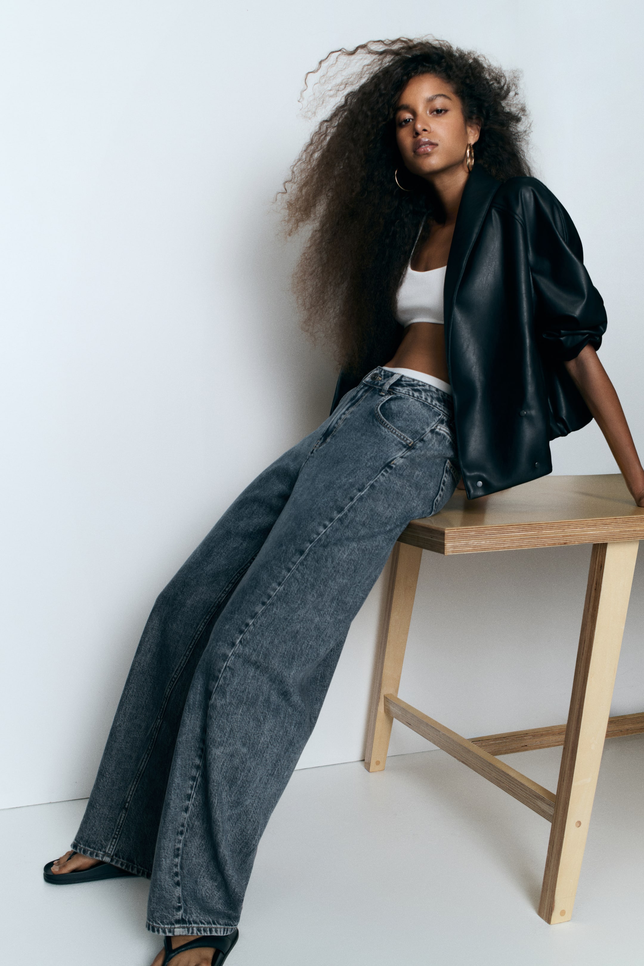 Vezi imaginea mai mare: Blugi din denim gri închis, cu talie înaltă și picioare curbate, voluminoase, sunt purtați de model. Blugii sunt asortați cu un top scurt alb și o jachetă neagră, purtată deschisă.