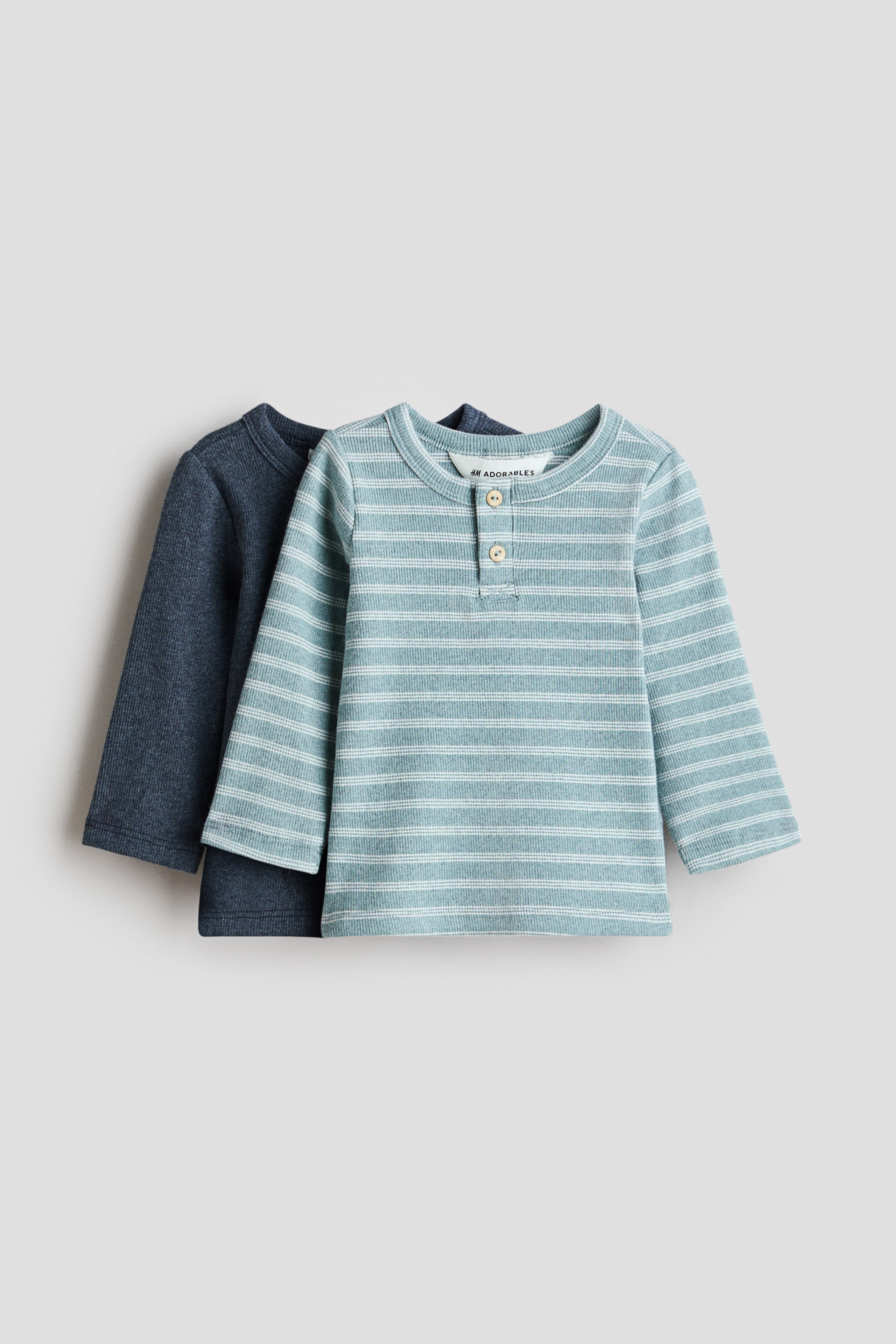查看大圖: 兩件H&M Adorables長袖羅紋針織上衣從正面展示。前景的上衣是淺藍色,帶有白色橫條紋,圓形羅紋領口和兩顆鈕扣門襟。直接在其後面是一件深藍色實色羅紋上衣。