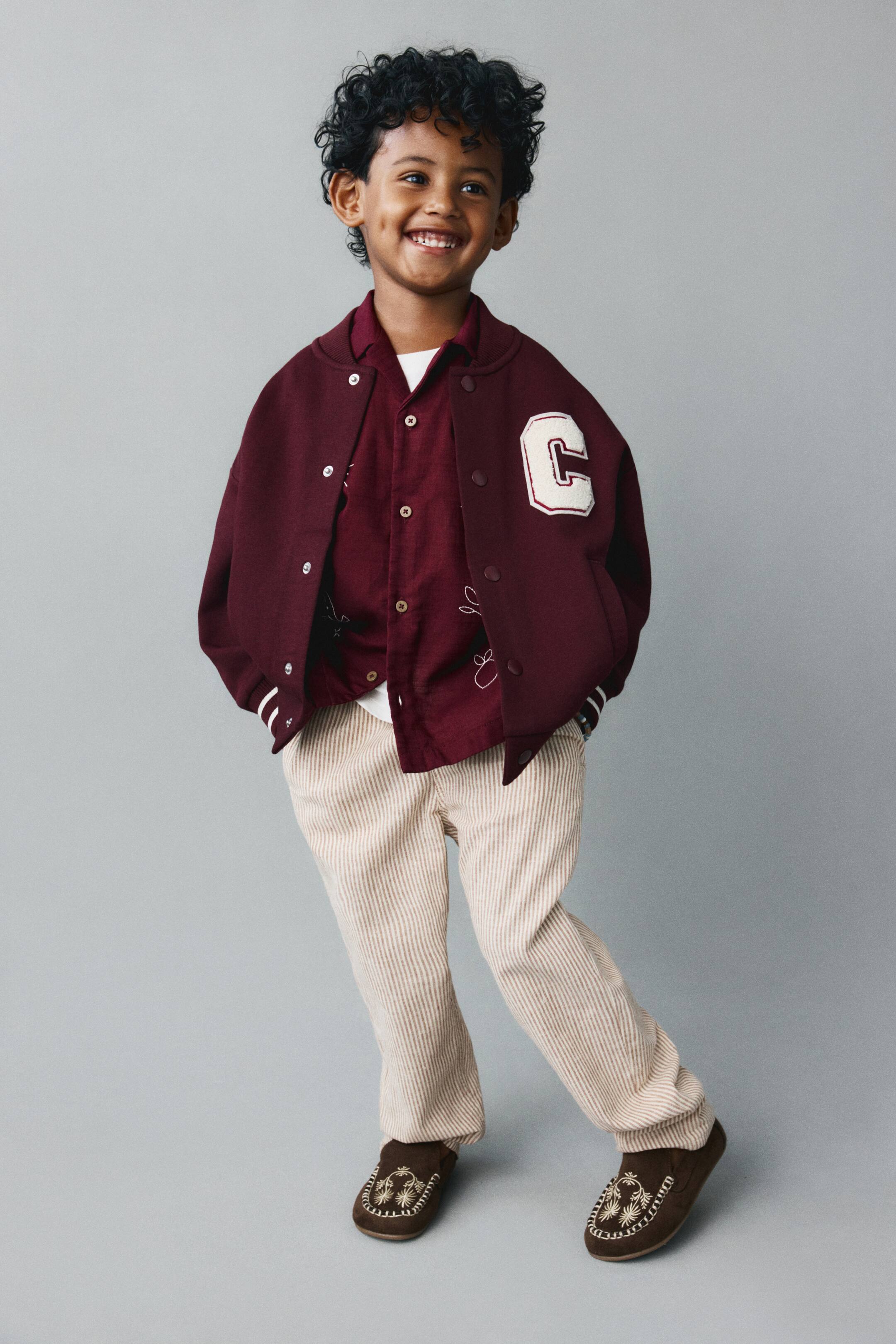 View larger image: A burgundy relaxed-fit baseball jacket is facing front, draped open over a matching button-down shirt to show its loose, comfortable silhouette. The jacket features a large white chenille "C" patch on the chest, a snap-button front, and ribbed cuffs with white stripe details.