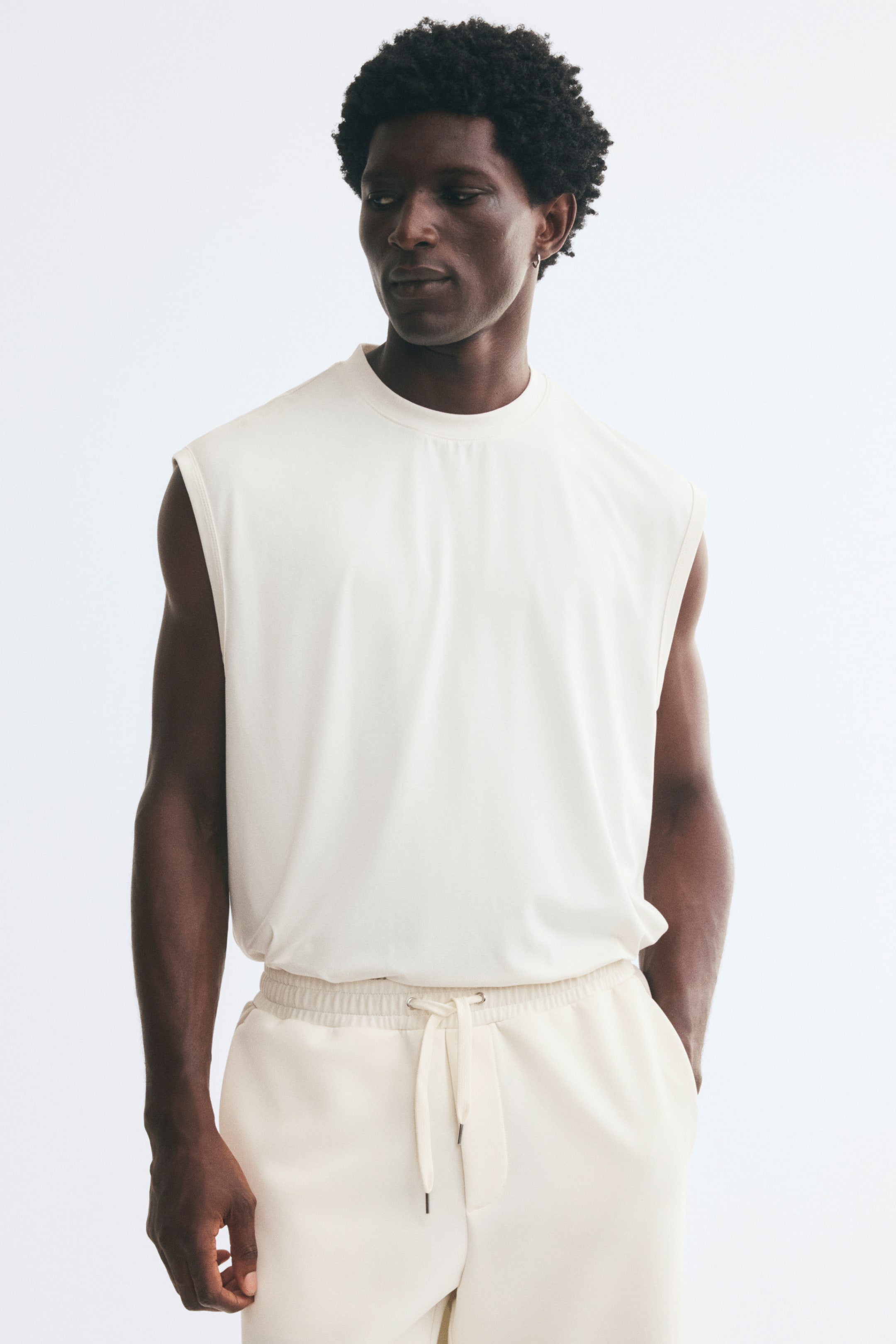 Voir une image plus grande: Le modèle porte un camisole blanc cassé à encolure ronde bordée de côtes et à profondes emmanchures. Le haut à coupe décontractée est rentré dans un pantalon de jogging blanc cassé assorti avec cordon de serrage.