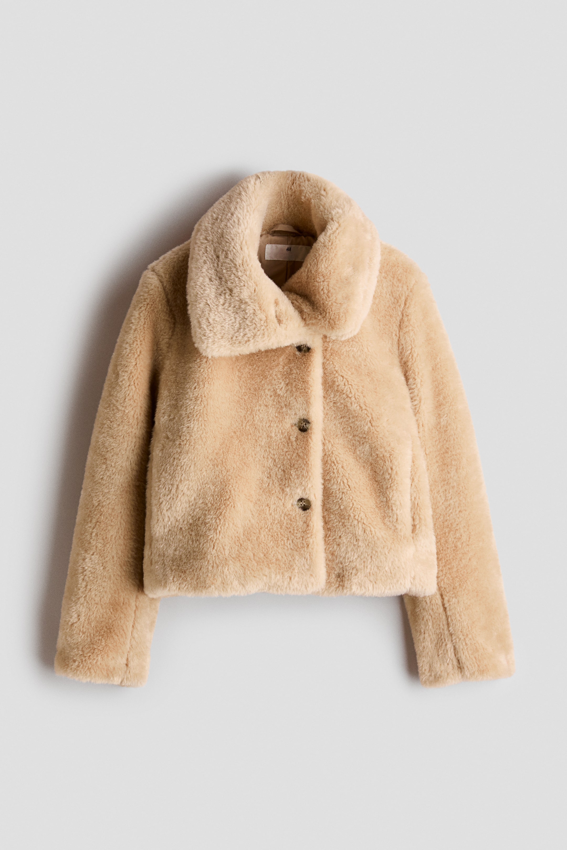 View larger image: A beige faux fur jacket is shown from the front, featuring a plush, wide collar, a cropped silhouette, and a three-button front closure. The jacket has long, straight sleeves and a soft, textured finish throughout.