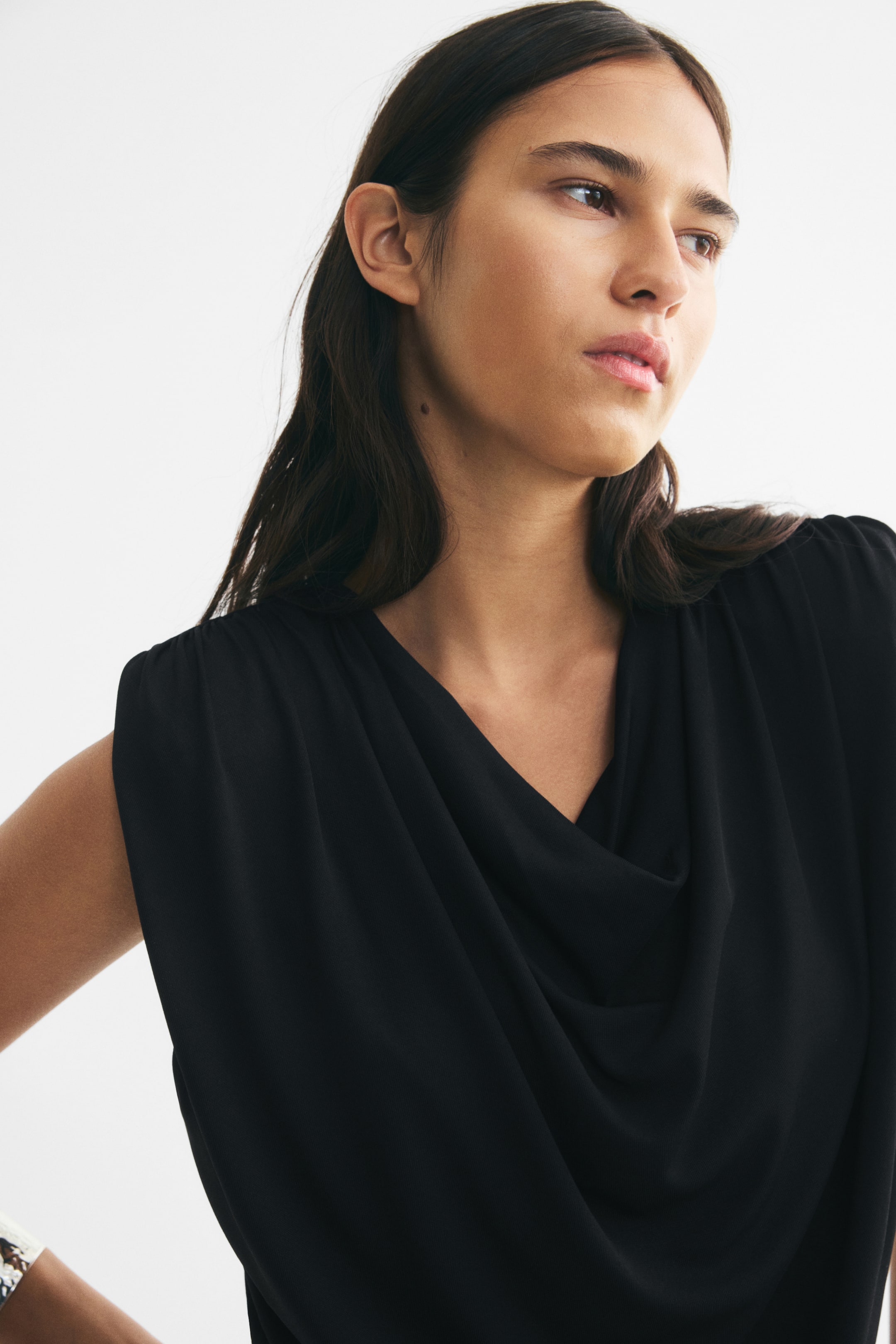 View larger image: A black, sleeveless top featuring a draped cowl neckline and gathered shoulders is being worn by the model. The top has structured shoulders that create a draped effect across the front.