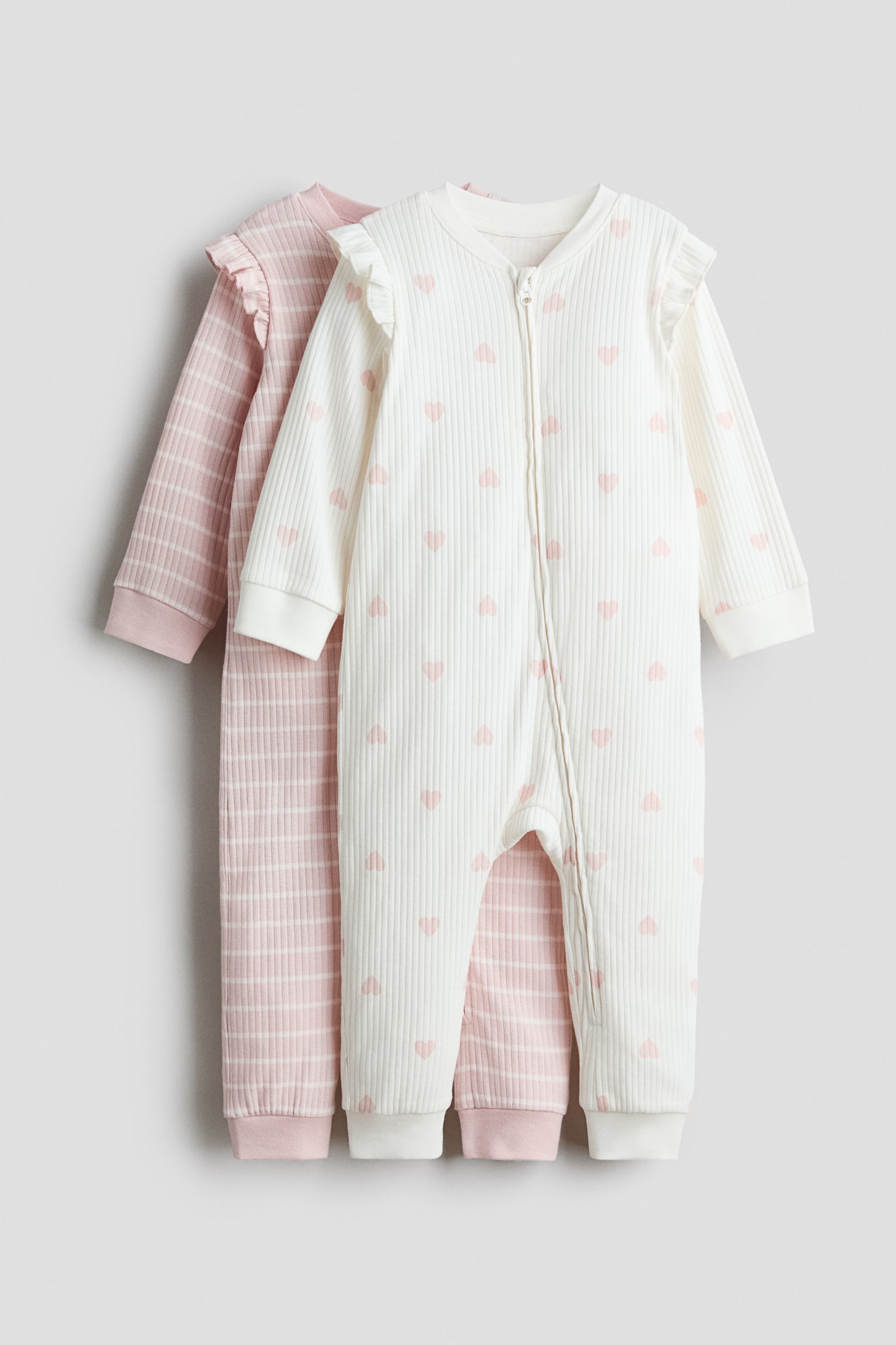 Two long-sleeve sleepsuits in ribbed cotton jersey are displayed from the front, featuring trimmed necklines, frills over the shoulders, and ribbed cuffs and hems. The front sleepsuit is off-white with a pink heart pattern and a zipper running down the front and along one leg, while the sleepsuit layered behind is light pink with white horizontal stripes.