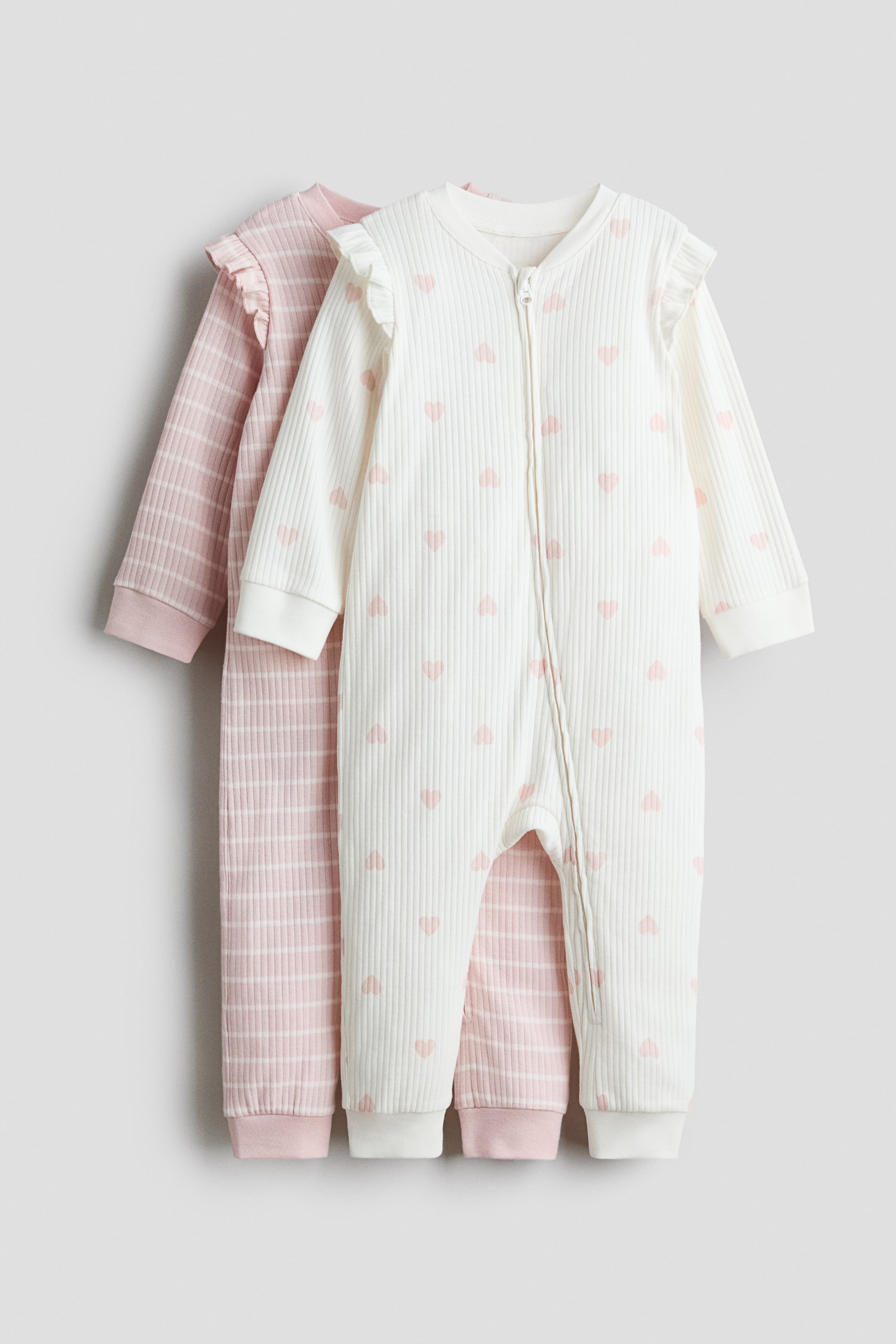 Grotere afbeelding bekijken: Twee sleepsuits met lange mouwen van ribgebreide katoenjersey zijn van voren afgebeeld, met afgewerkte halslijnen, ruches over de schouders en ribgebreide boorden en zomen. De sleepsuit op de voorgrond is gebroken wit met een roze hartjespatroon en heeft een rits aan de voorkant en langs één pijp, terwijl de sleepsuit daarachter lichtroze is met witte horizontale strepen.