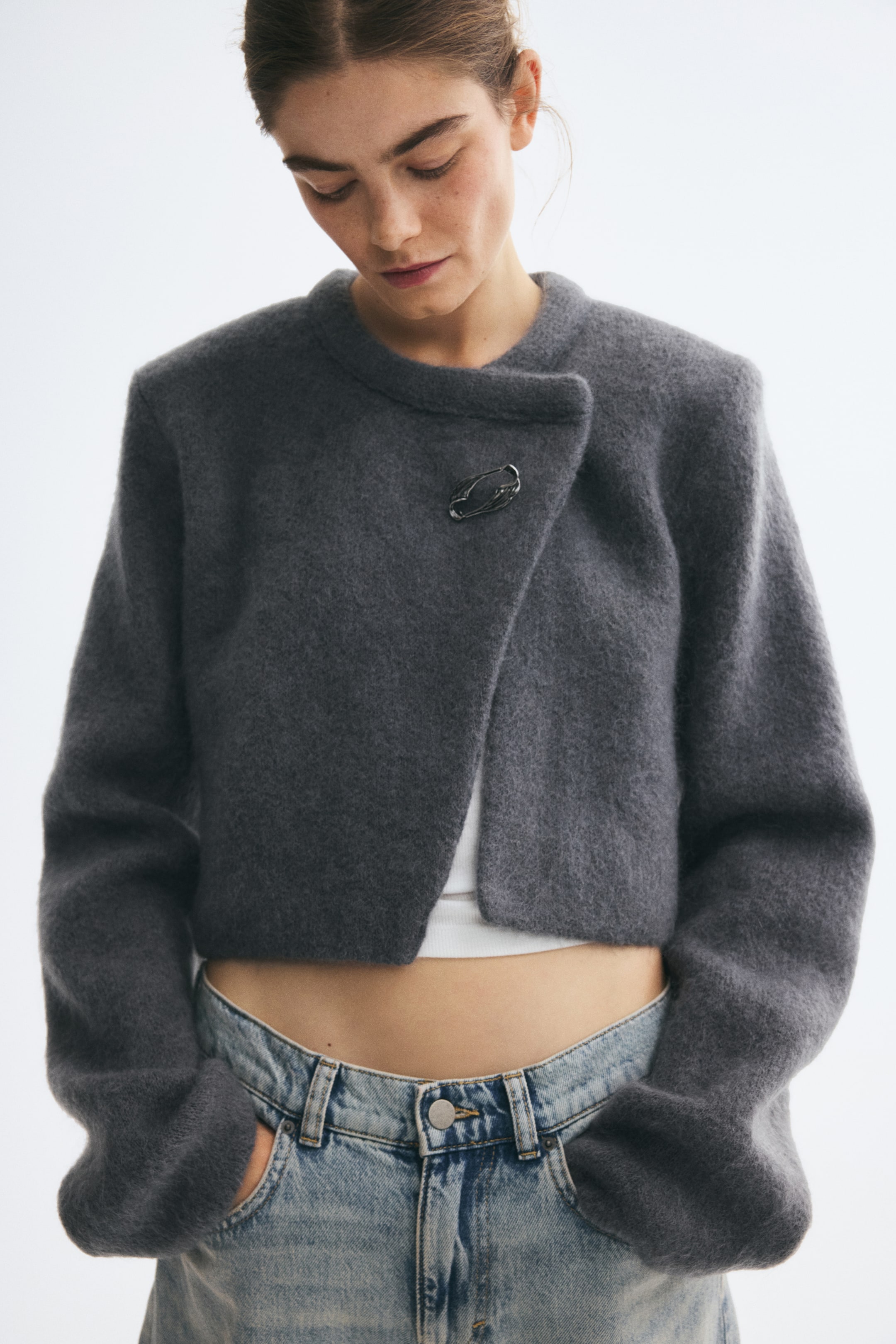 Vezi imaginea mai mare: Un cardigan gri scurt, dintr-un amestec moale de mohair și lână, este purtat de model, suprapus peste un top alb și o pereche de jeans deschiși la culoare. Cardiganul are decolteu rotund, umeri structurați, mâneci lungi și o închidere frontală asimetrică, fixată cu un ac abstract în nuanță argintie.